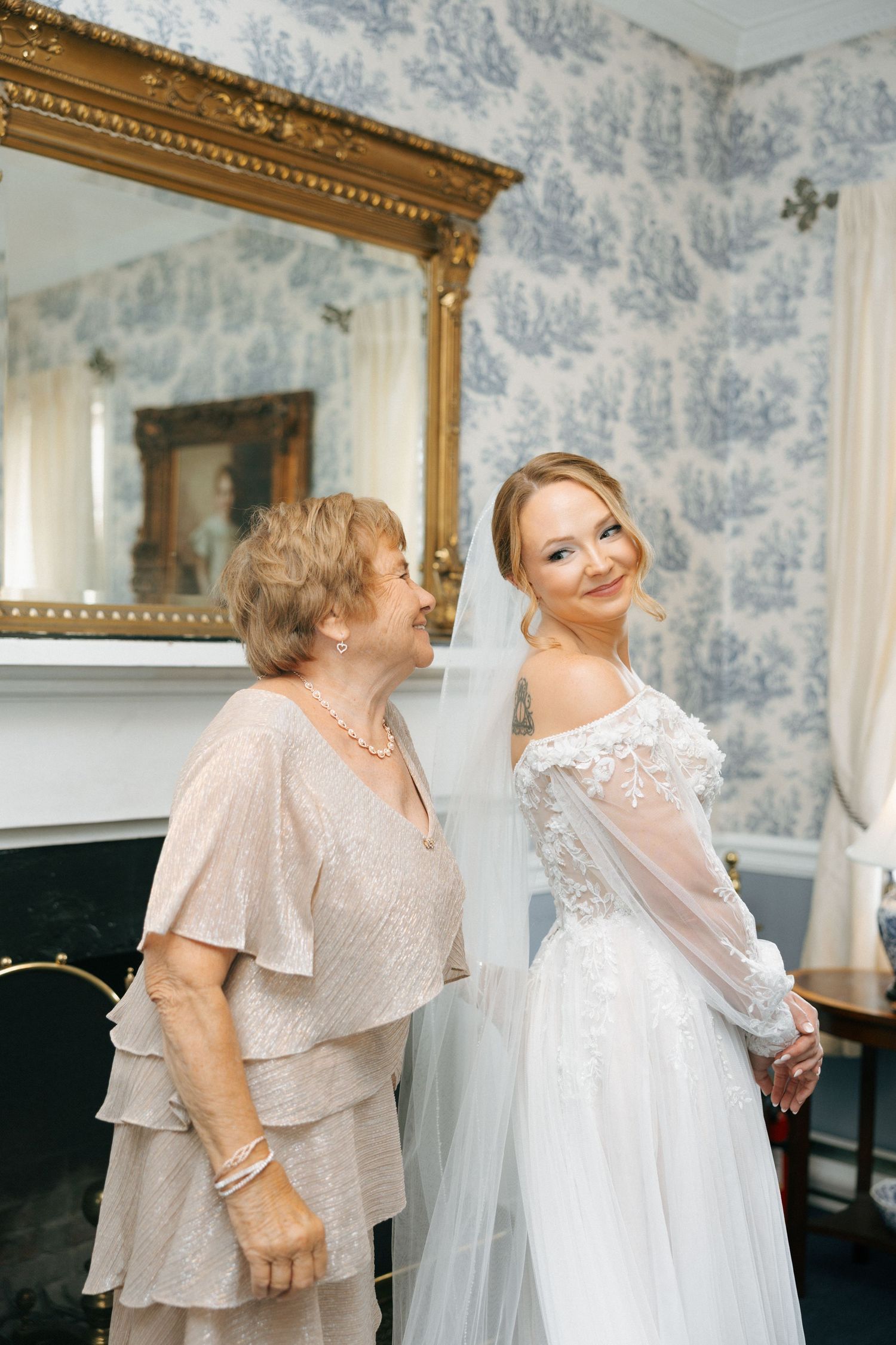 Two people share an emotional moment in an elegant room with blue and white toile wallpaper and gold-framed mirrors.