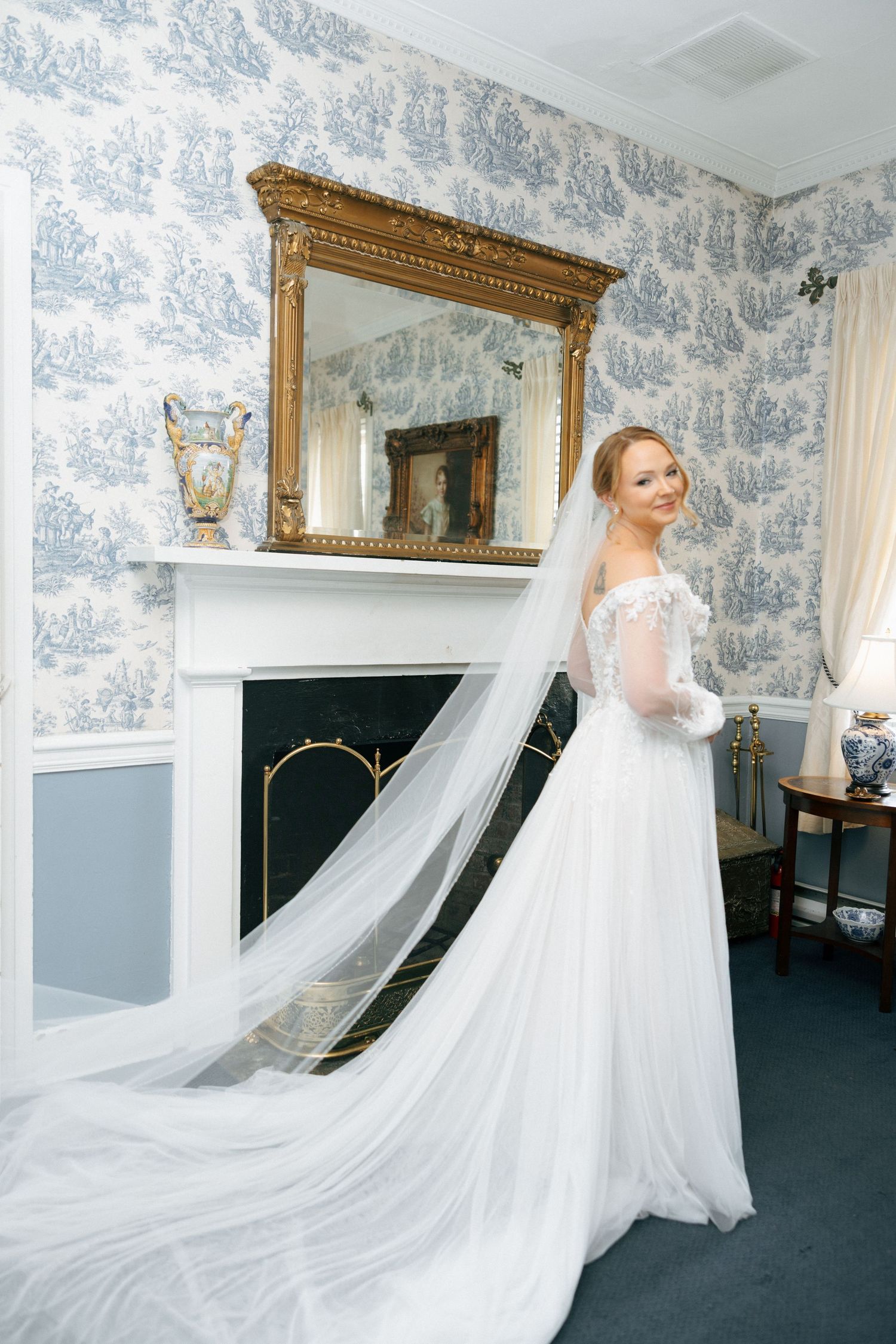 A flowing white wedding dress with cathedral veil is showcased in a blue and white toile wallpapered room.