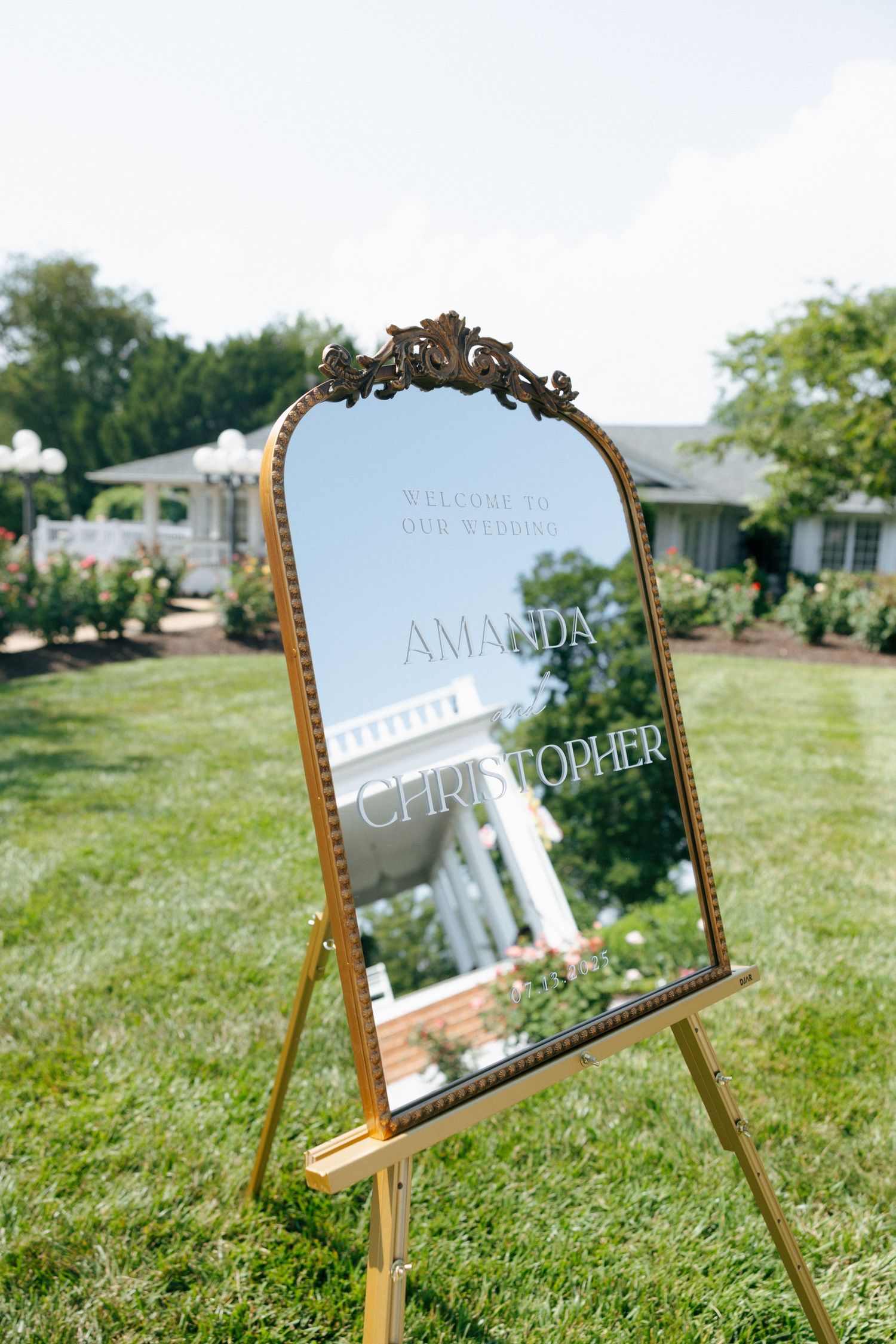 Ornate gold-framed mirror wedding welcome sign displayed on wooden easel in garden setting.