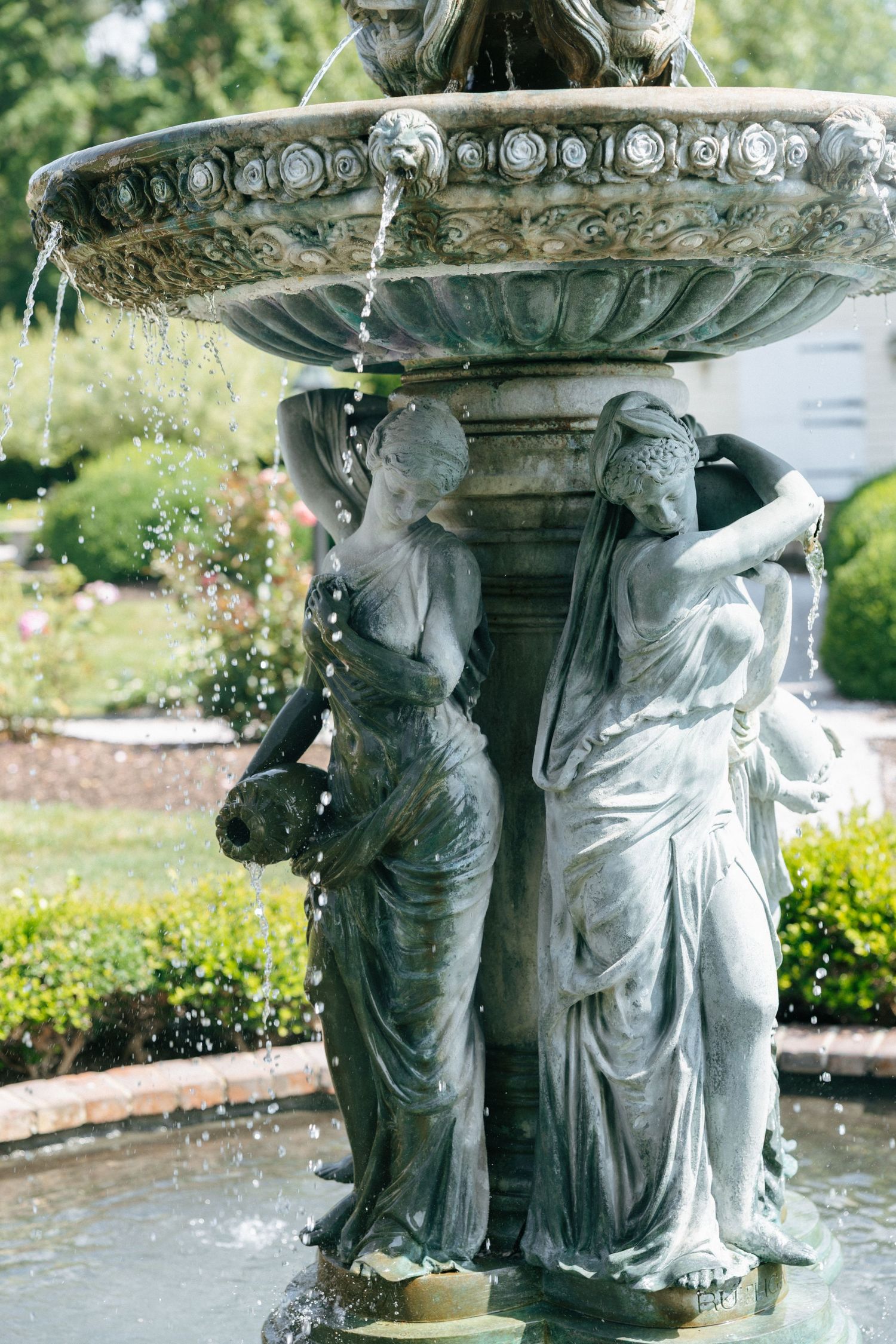 Ornate garden fountain with classical statues supporting a decorative bowl with flowing water in a lush garden setting.