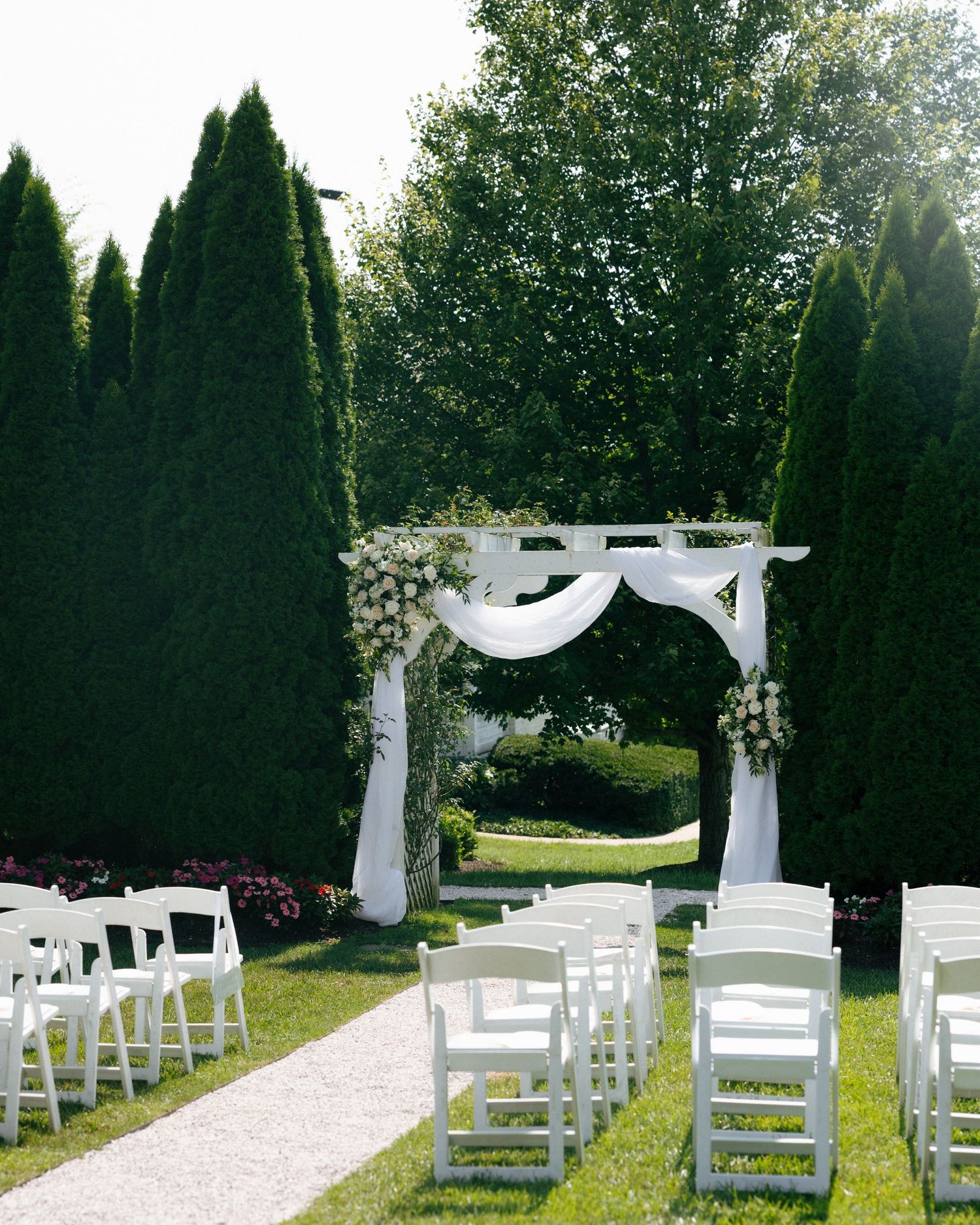 White wedding ceremony setup with chairs and fabric draped arches in an outdoor garden setting with tall evergreen trees.