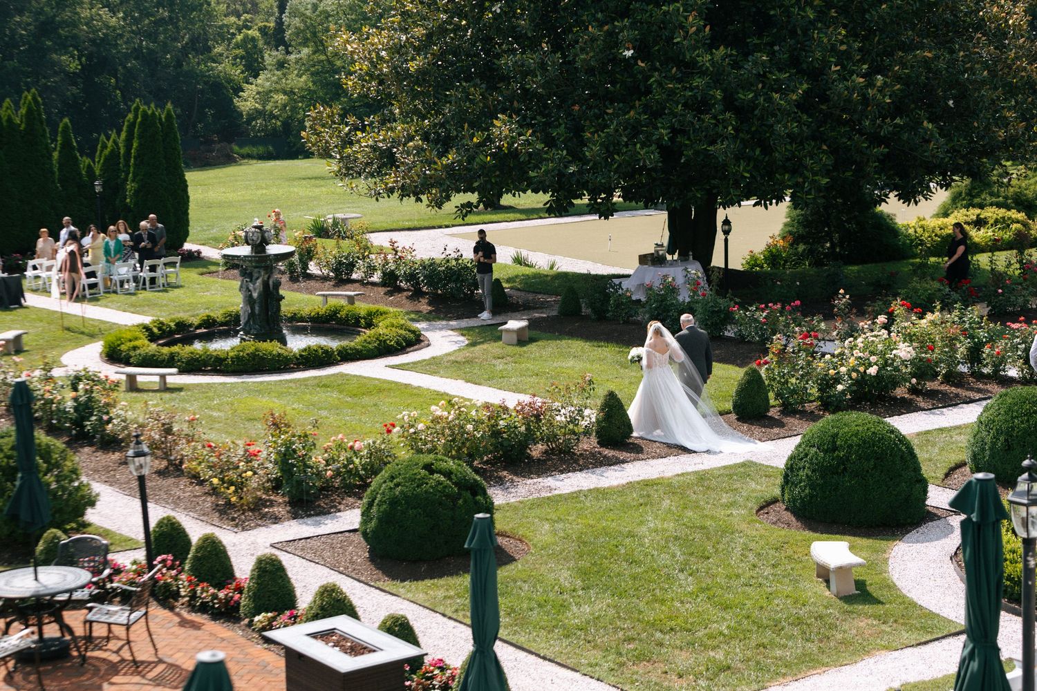 Aerial view of a formal garden wedding ceremony with guests gathered around curved pathways and manicured lawns.