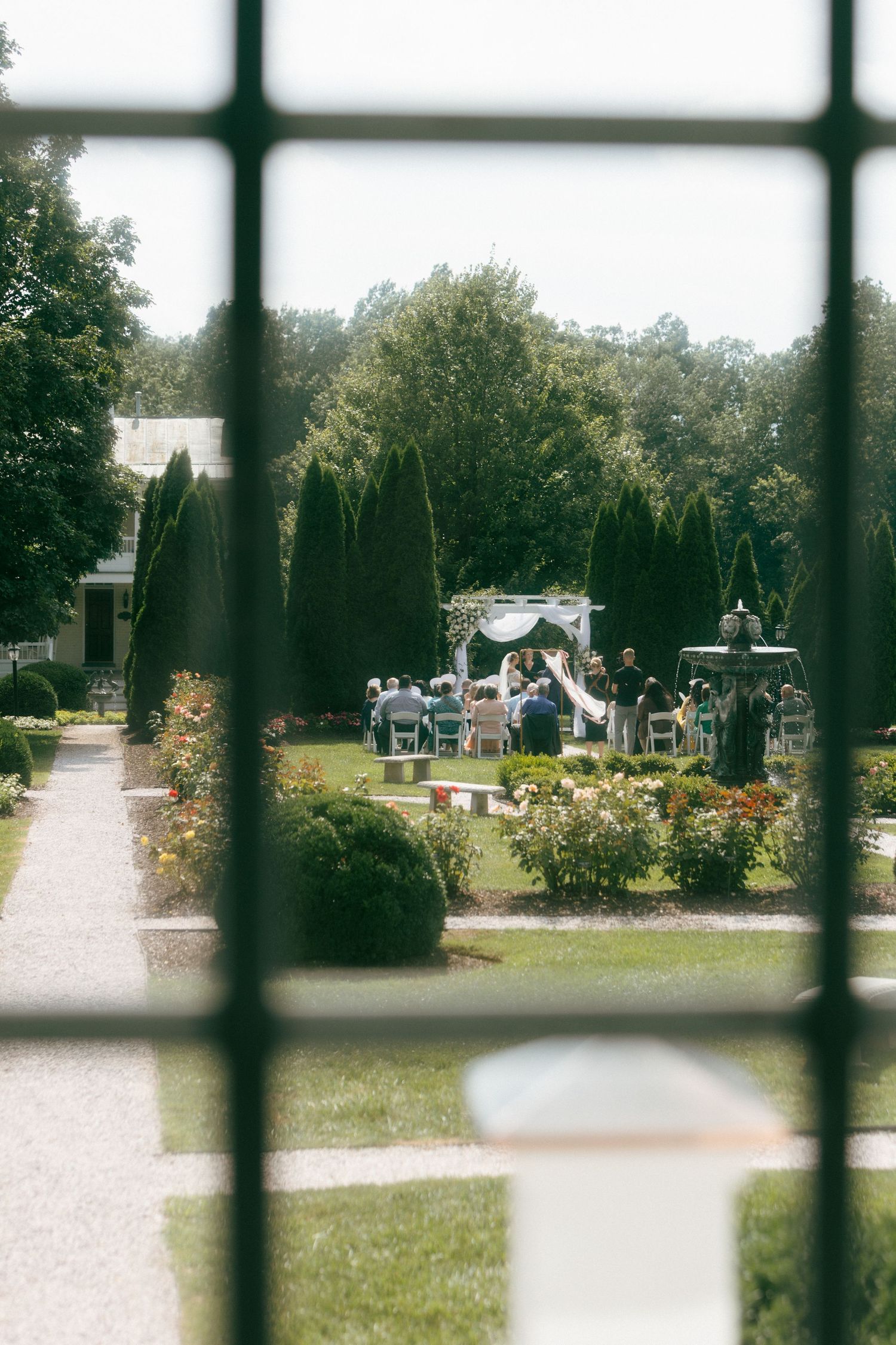 A wedding ceremony setup viewed through window panes with a white gazebo and guests in a garden surrounded by evergreens.