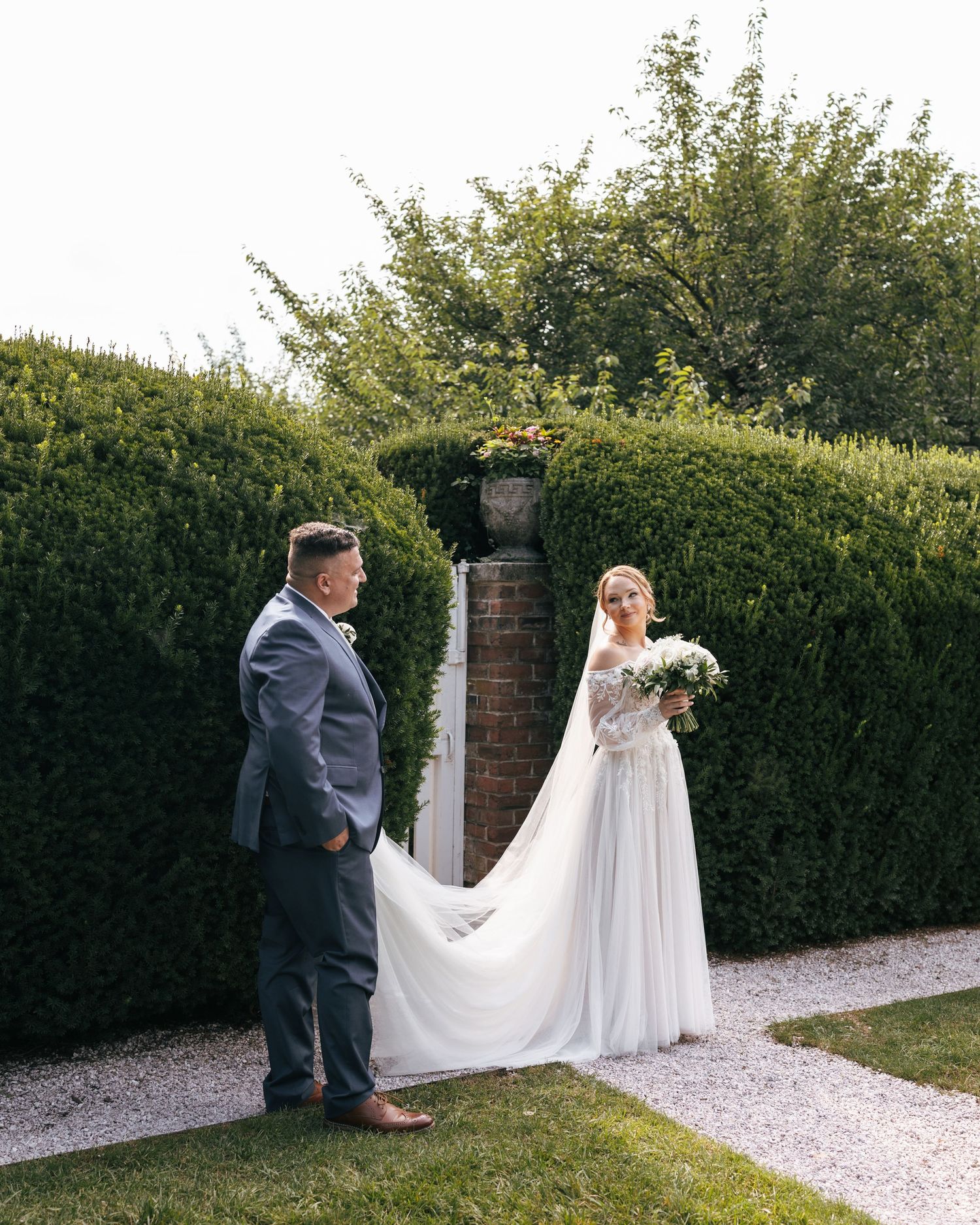 Wedding couple walking together on a gravel path lined with manicured hedges in a garden setting.