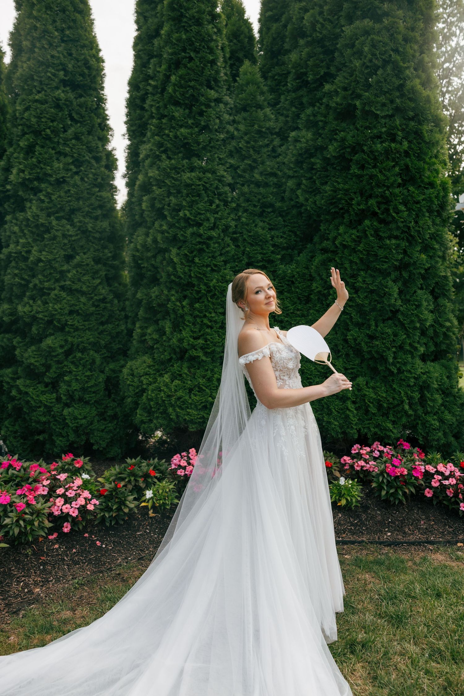 A flowing white wedding gown with long veil against a backdrop of tall evergreen trees and pink flowers.