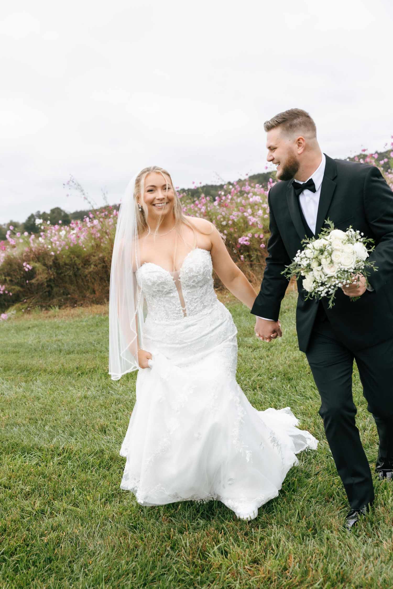 A bride and groom walk together on grassy hillside with pink wildflowers blooming in the background.