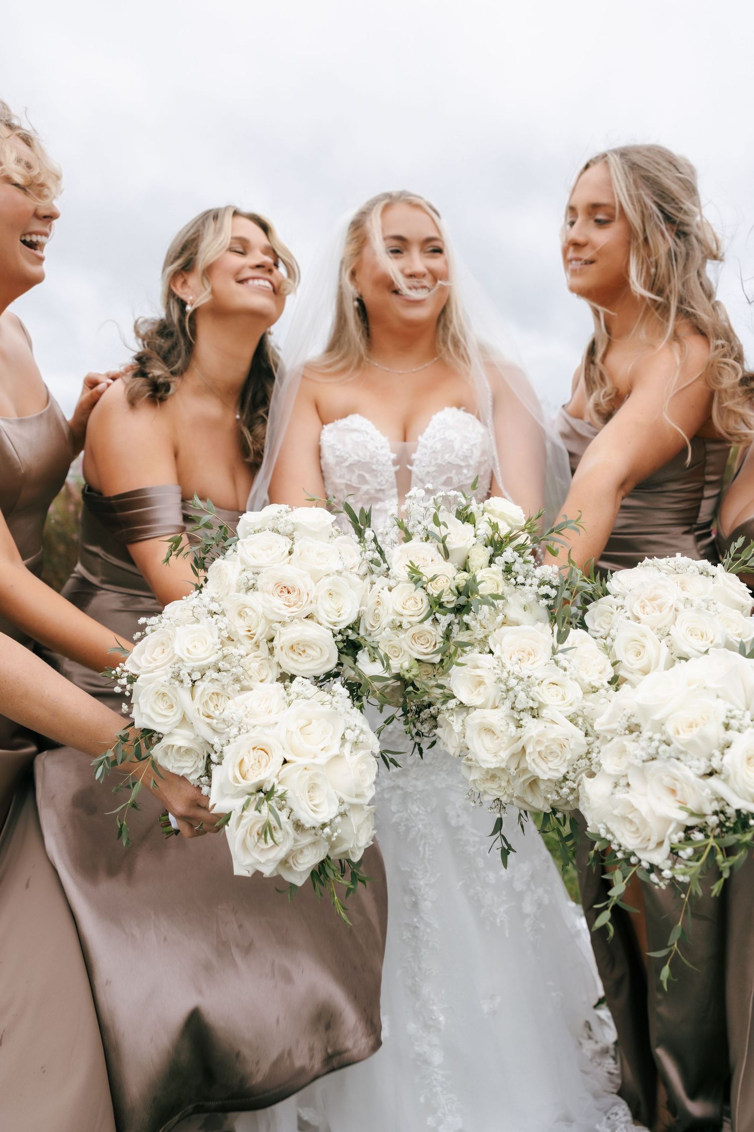 A bridal party in taupe dresses holds elegant white and cream floral bouquets while sharing joyful laughter.