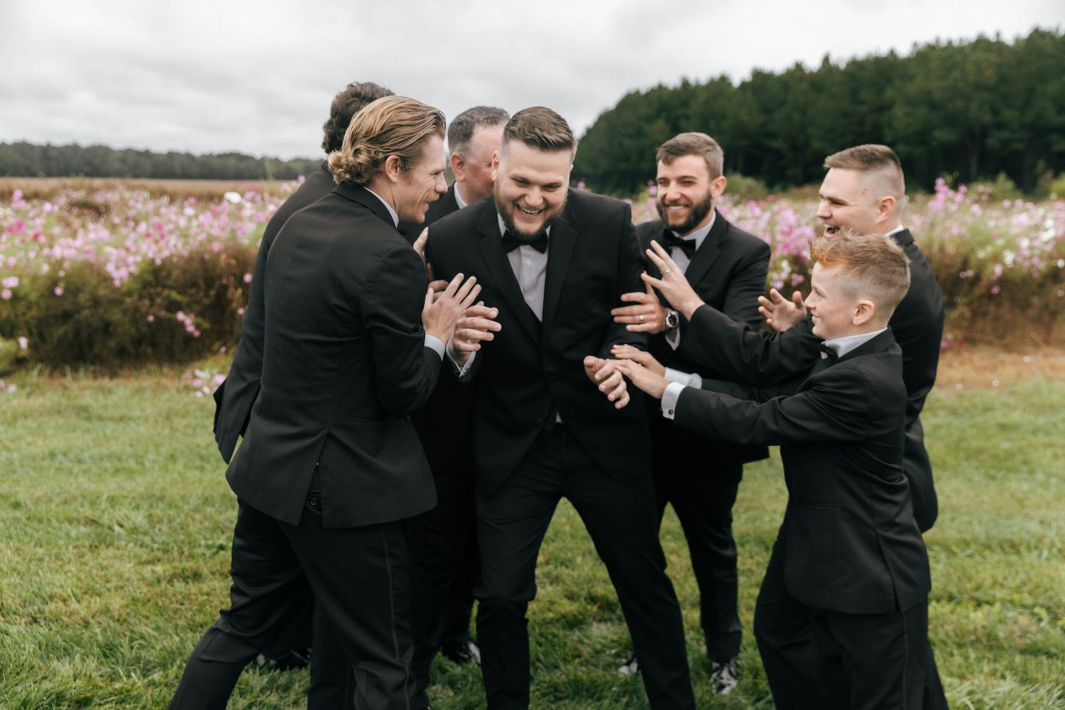 Groomsmen in black suits share a celebratory drink together in an outdoor field setting.