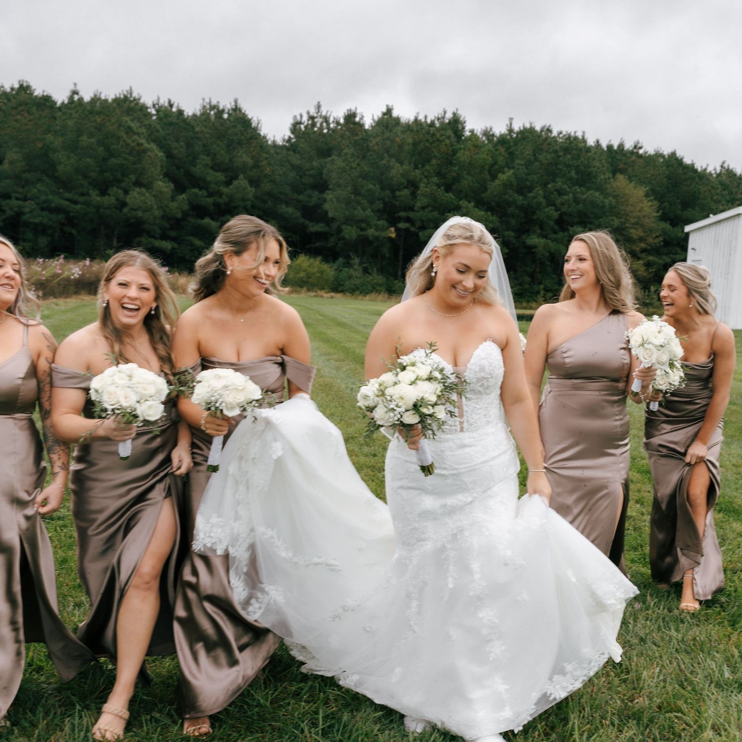 Bridesmaids in taupe dresses help bride with her flowing white gown in an outdoor field setting.