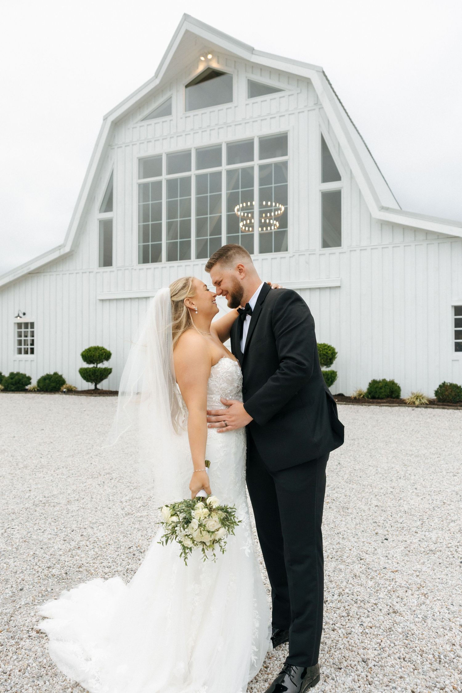 A romantic wedding couple shares an intimate moment outside a white barn venue on a gravel driveway.