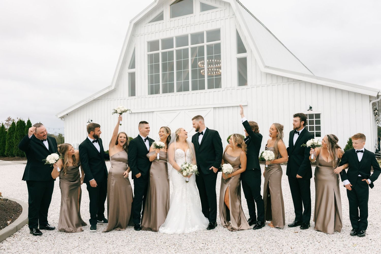 Wedding party in formal attire poses in front of white church building on a cloudy day.