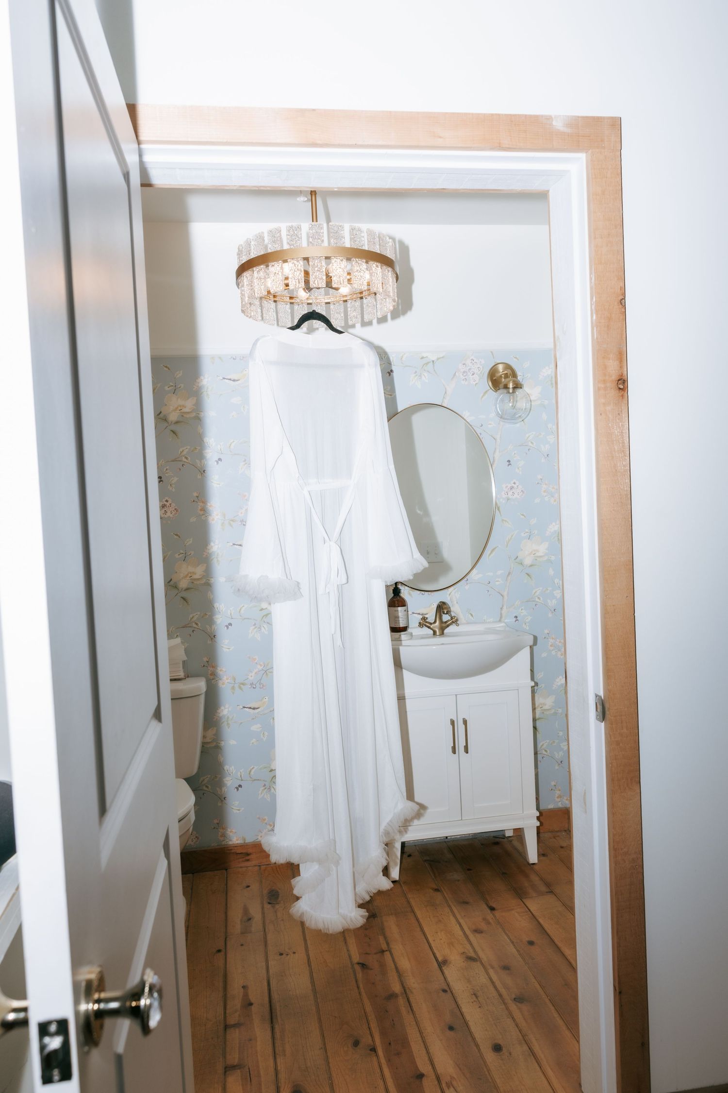Elegant bridal dressing room with hardwood floors, white gown display, and crystal chandelier in vintage style setting.