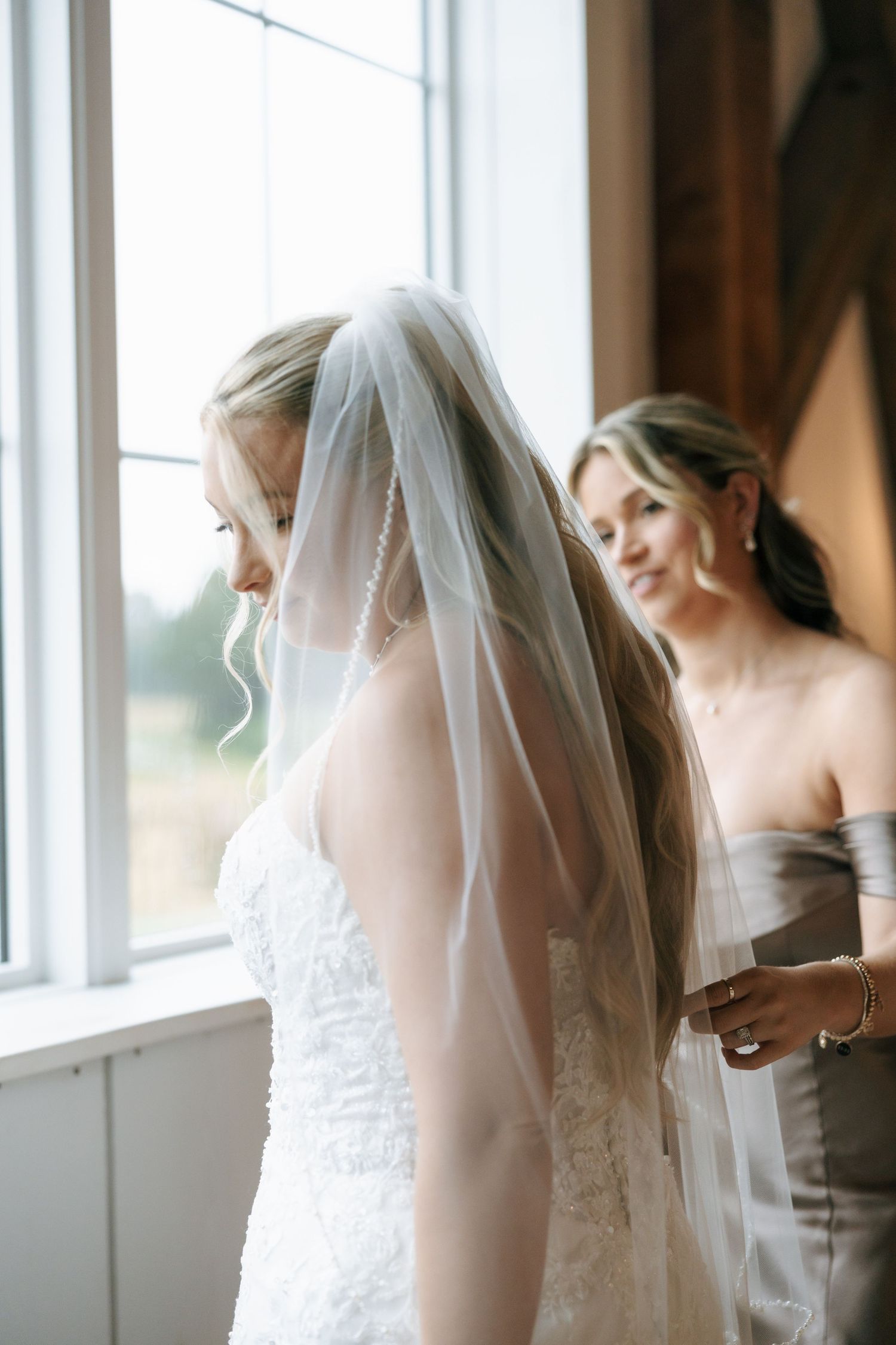 A bride in a white wedding dress and veil stands by large windows in an elegant indoor setting.