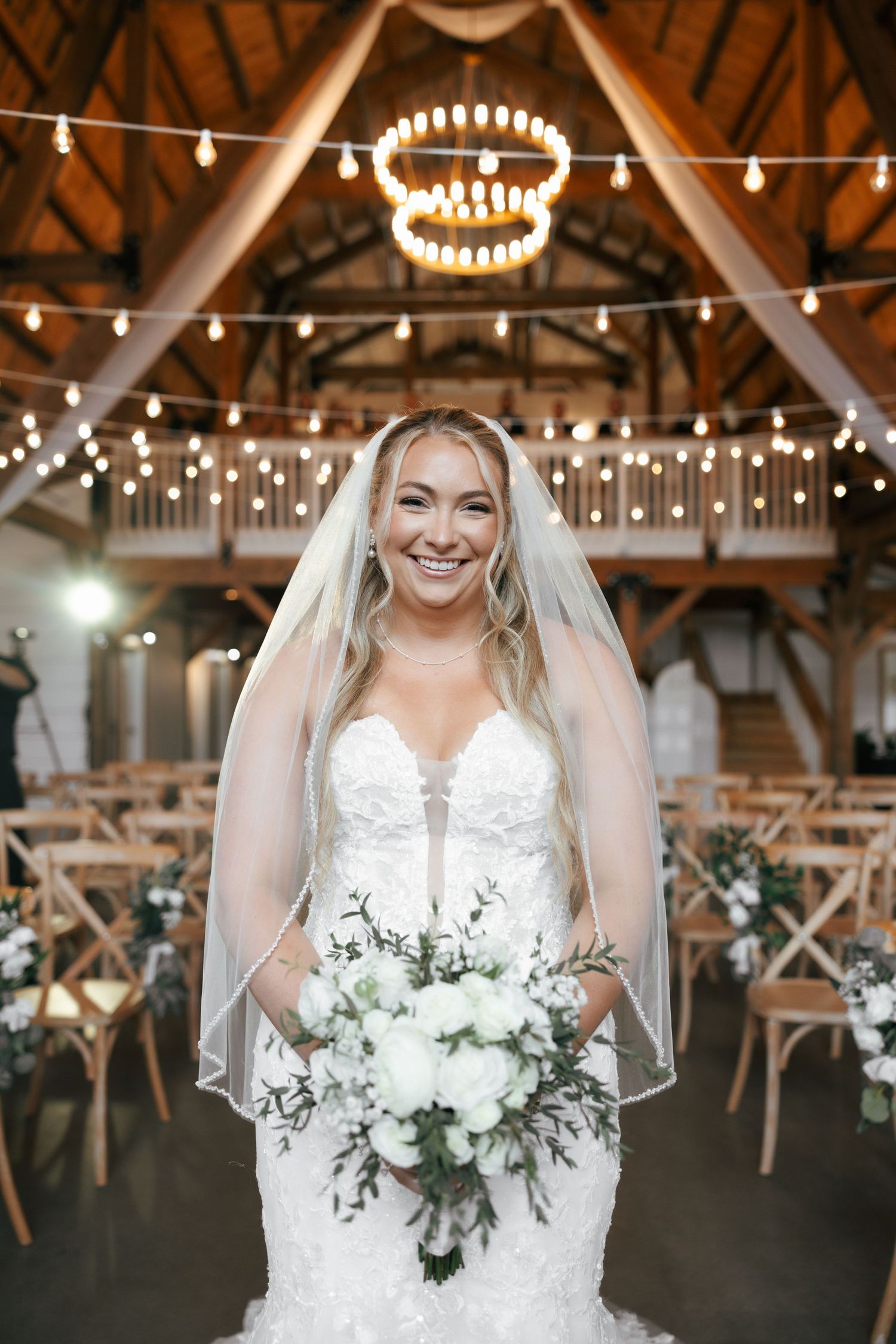 A bride in a white lace wedding dress holds a white and green floral bouquet in an elegant rustic barn venue.