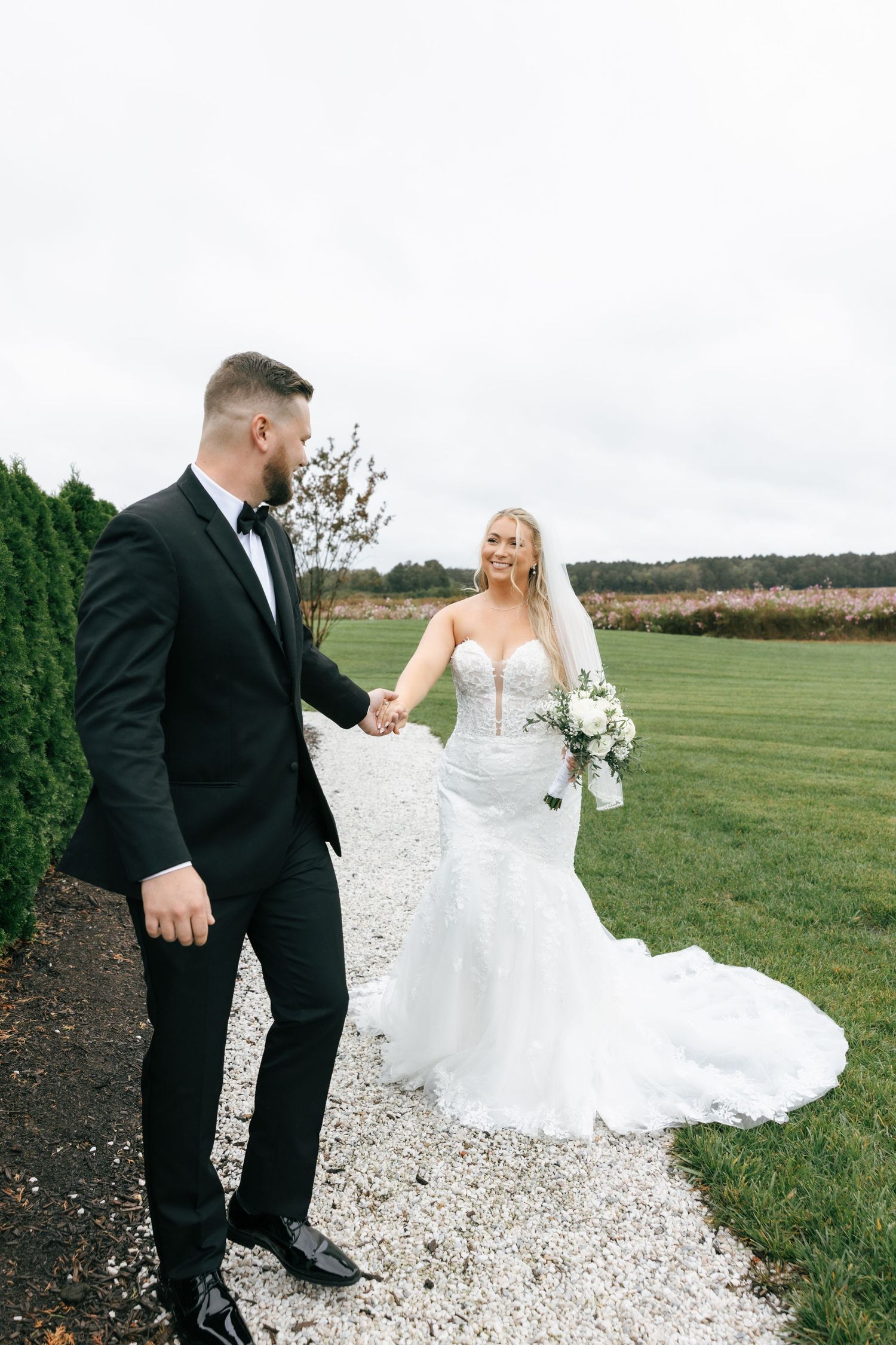 Wedding couple walking on gravel path outdoors with green fields in background.