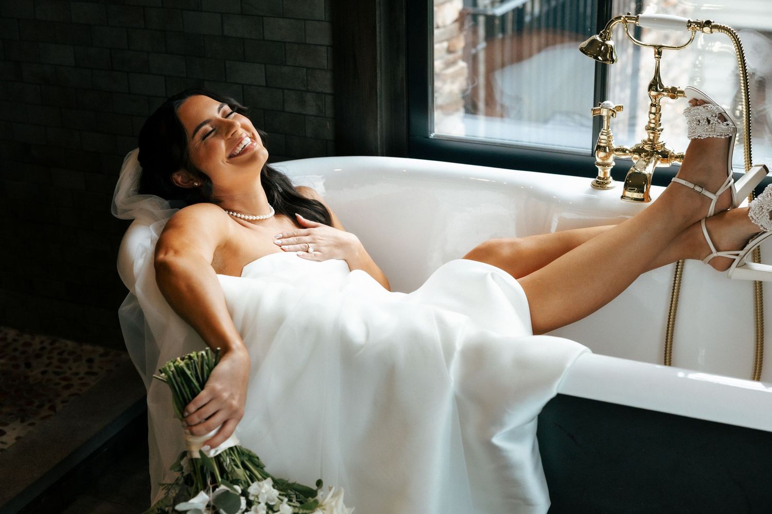 Smiling bride in white wedding dress relaxing in a freestanding bathtub holding flowers wearing heels.