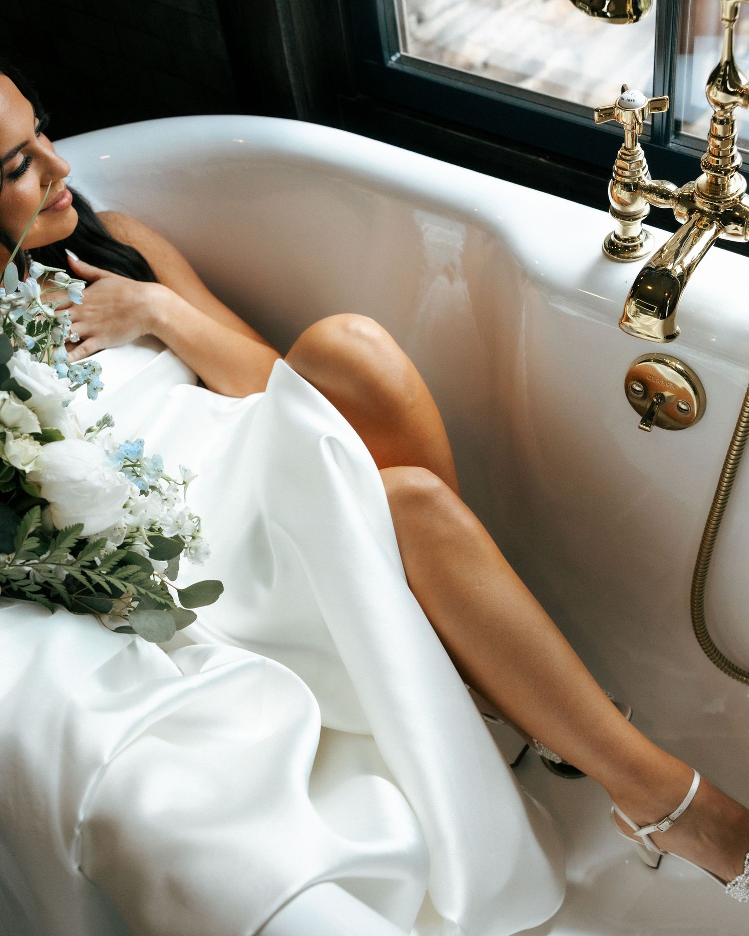 Bride in white gown holding blue floral bouquet, reclining elegantly in a vintage clawfoot bathtub with gold fixtures.