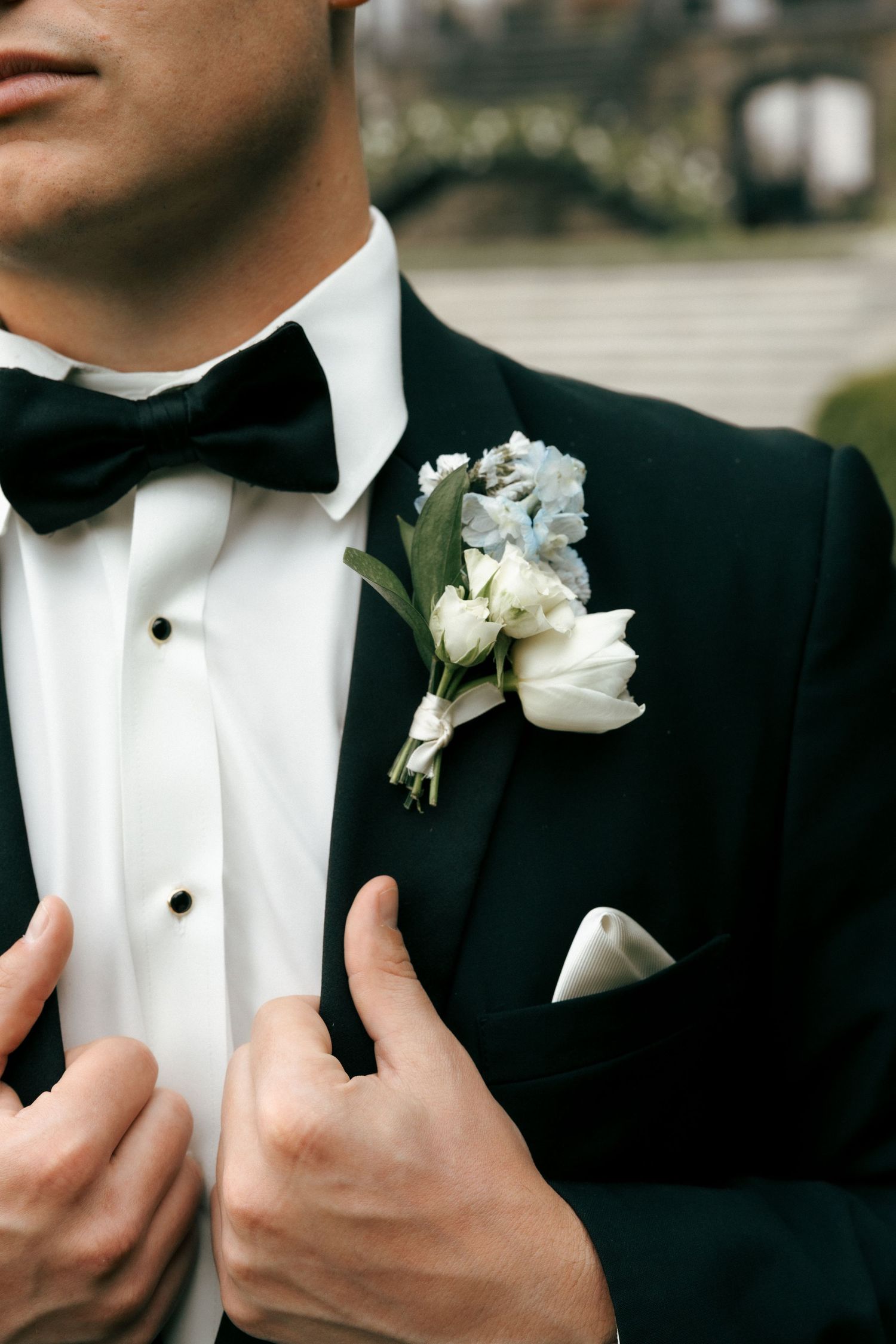 Groom in black tuxedo with bow tie adjusting boutonniere of white roses and blue flowers on lapel.