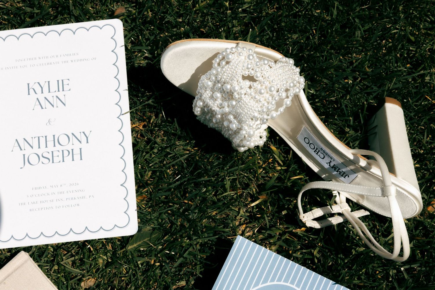 White pearl-embellished wedding shoes beside a Kylie Ann and Anthony Joseph wedding invitation on green grass.