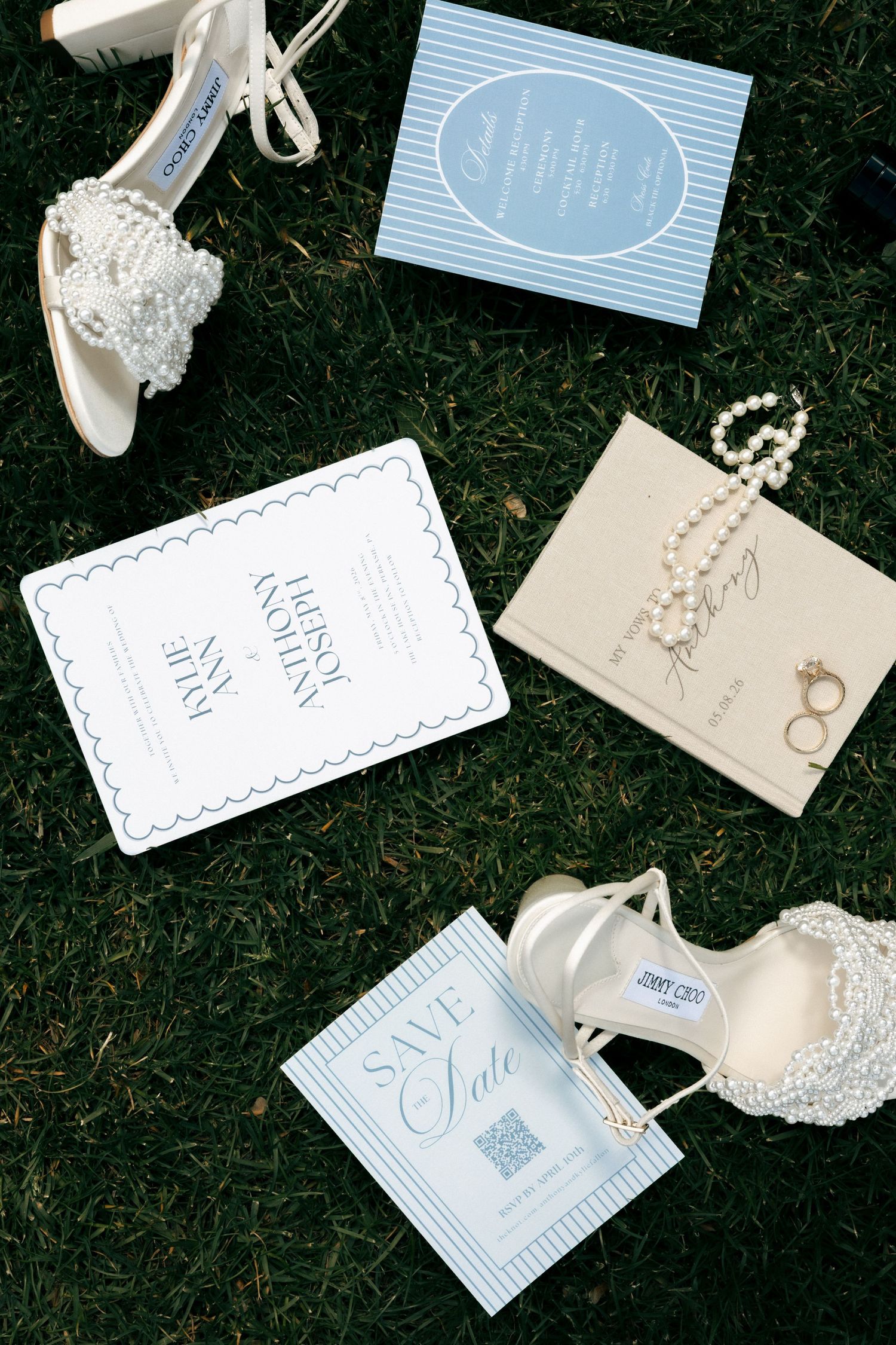 Wedding flat lay with pearl heels, blue stationery, save the date cards, pearl necklace, and gold rings on green grass.