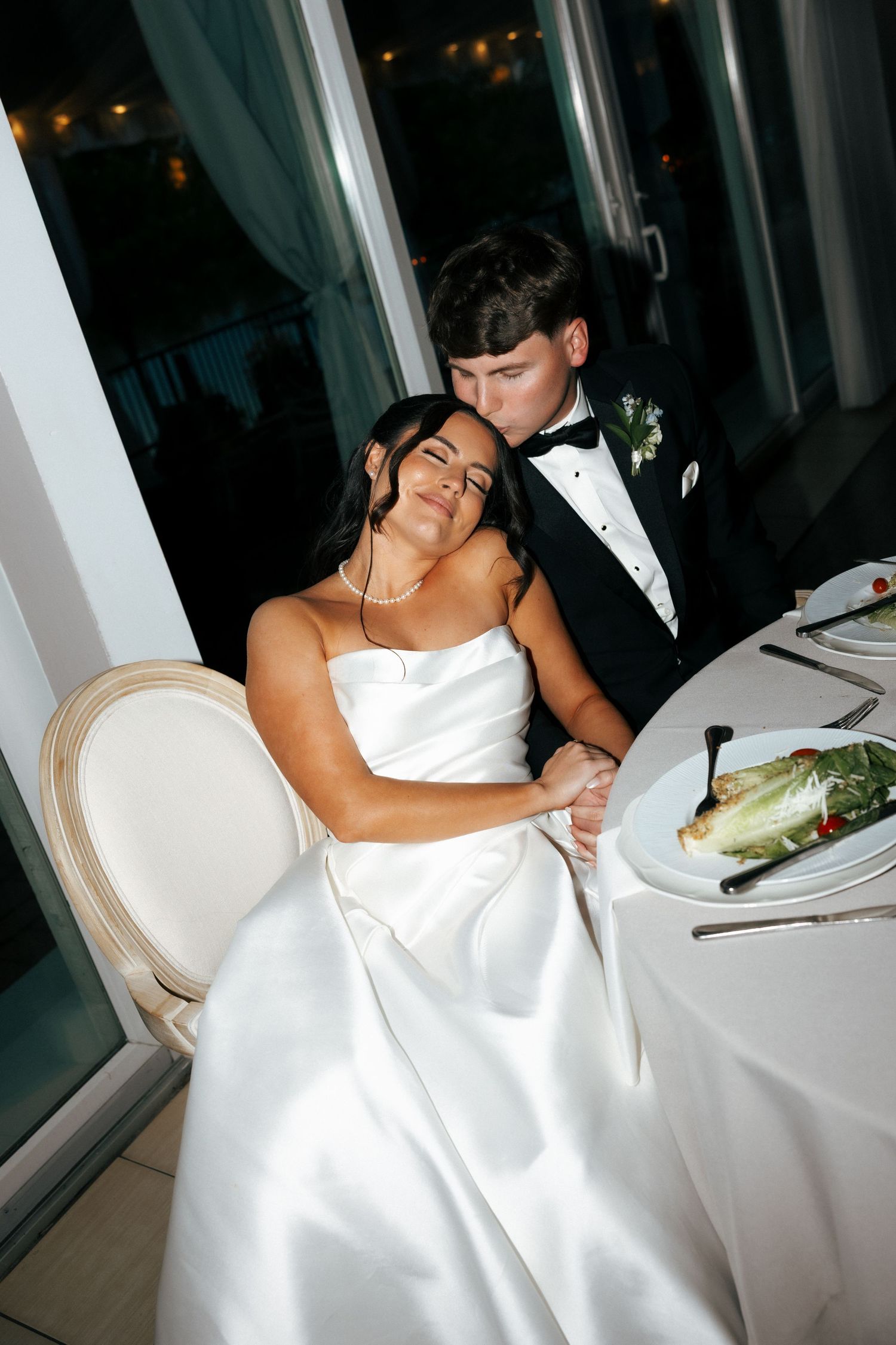 Bride in white satin gown seated at reception table while groom in tuxedo kisses her cheek tenderly.