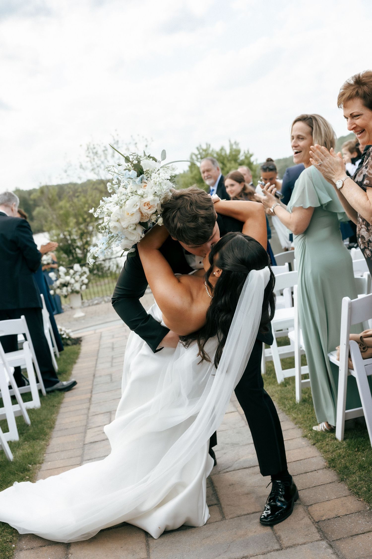 Bride and groom share a romantic dip kiss at outdoor wedding ceremony while guests applaud in background.