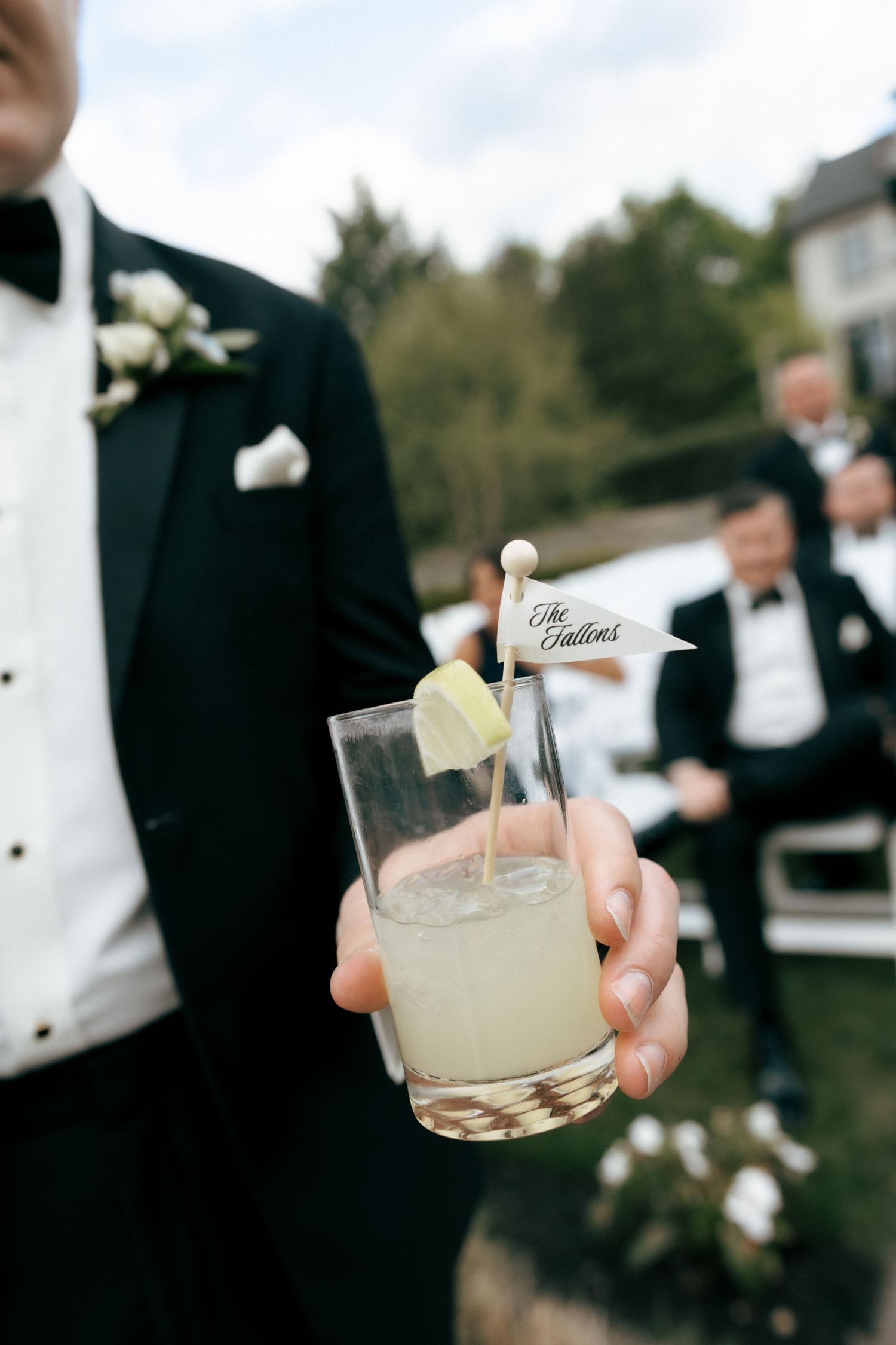 Groom in tuxedo holding signature cocktail with personalized flag pick at outdoor wedding reception.