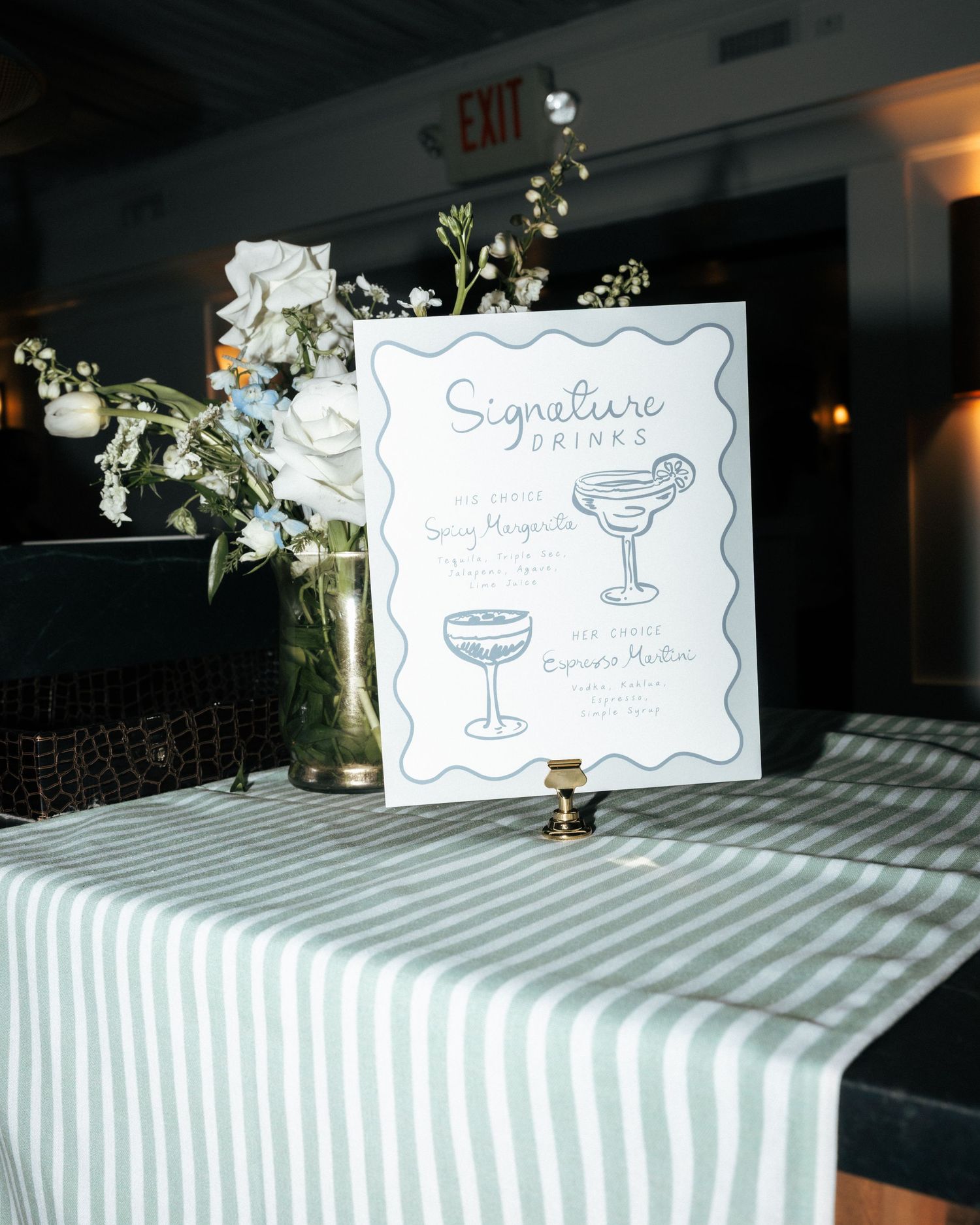 Elegant wedding signature drinks sign displayed beside white floral arrangement on striped linen table.