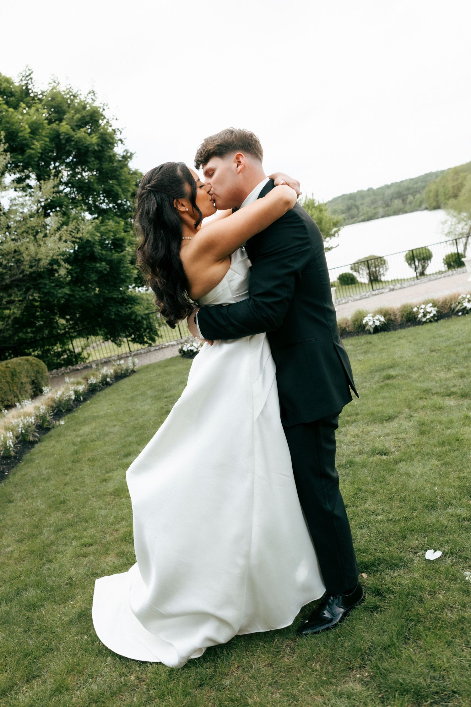 A bride and groom share a romantic kiss while embracing on a green lawn with trees and a lake in the background.