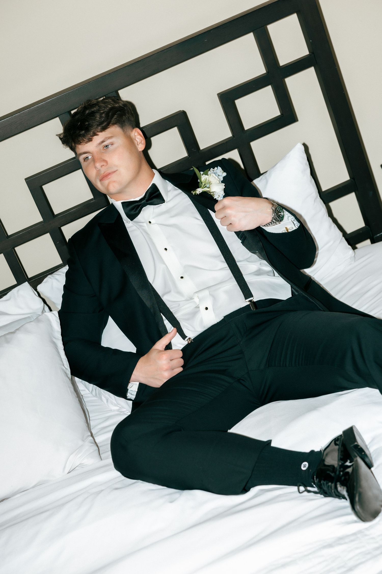 Young man in black tuxedo with bow tie and boutonniere poses on white bed, wearing dress shoes and a watch.