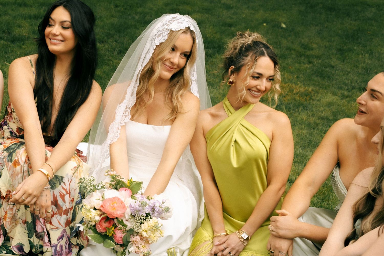 Bride in white dress and veil sits with bridesmaids in colorful gowns on green grass holding floral bouquet.