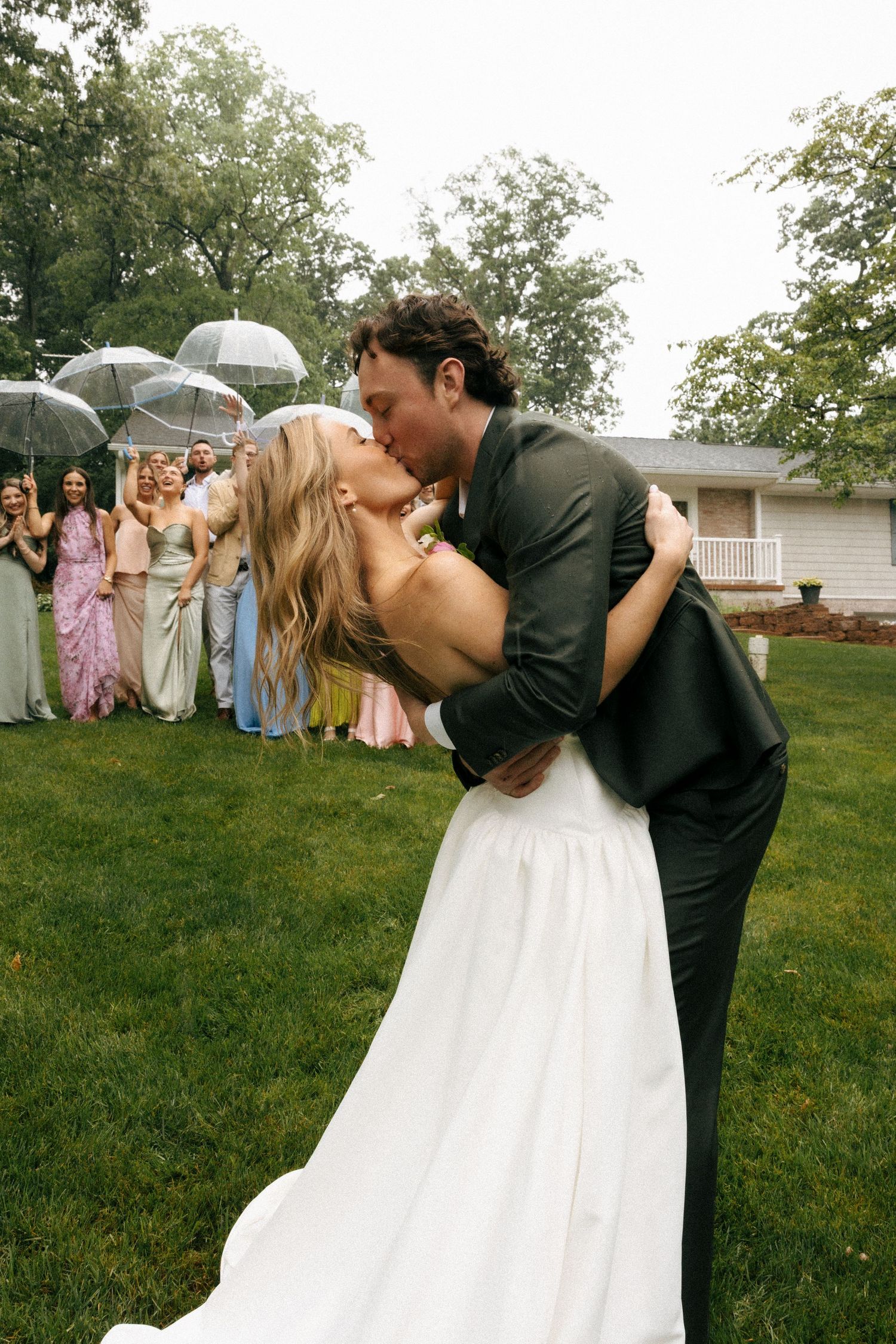 Bride and groom share a romantic kiss on green lawn while wedding guests hold clear umbrellas in background.