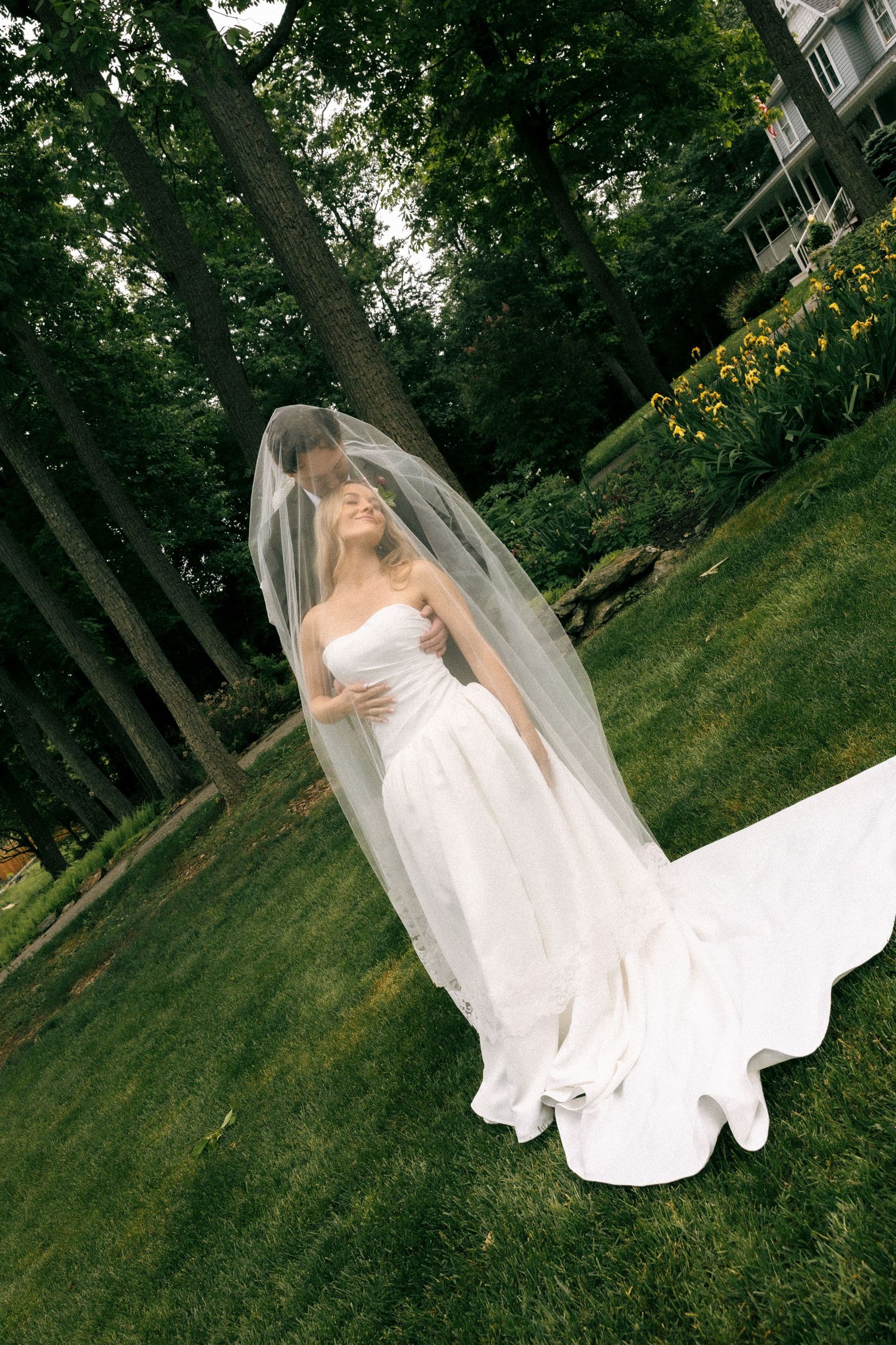 Bride in strapless white gown with flowing veil and train poses outdoors on green lawn surrounded by trees.