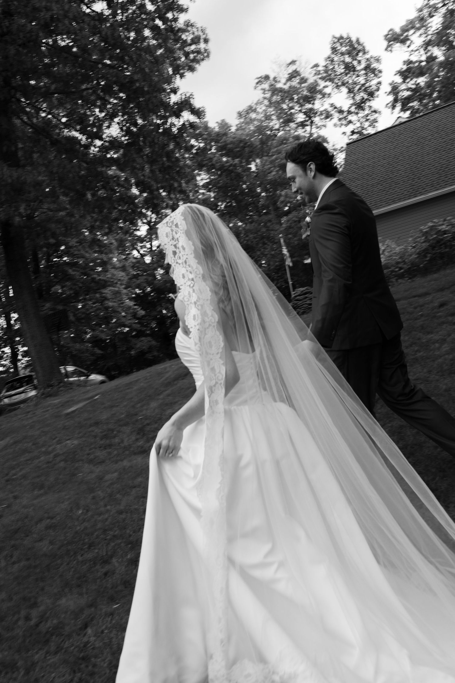 Black and white photo of a bride in a lace wedding gown and veil walking with a groom in a suit outdoors.