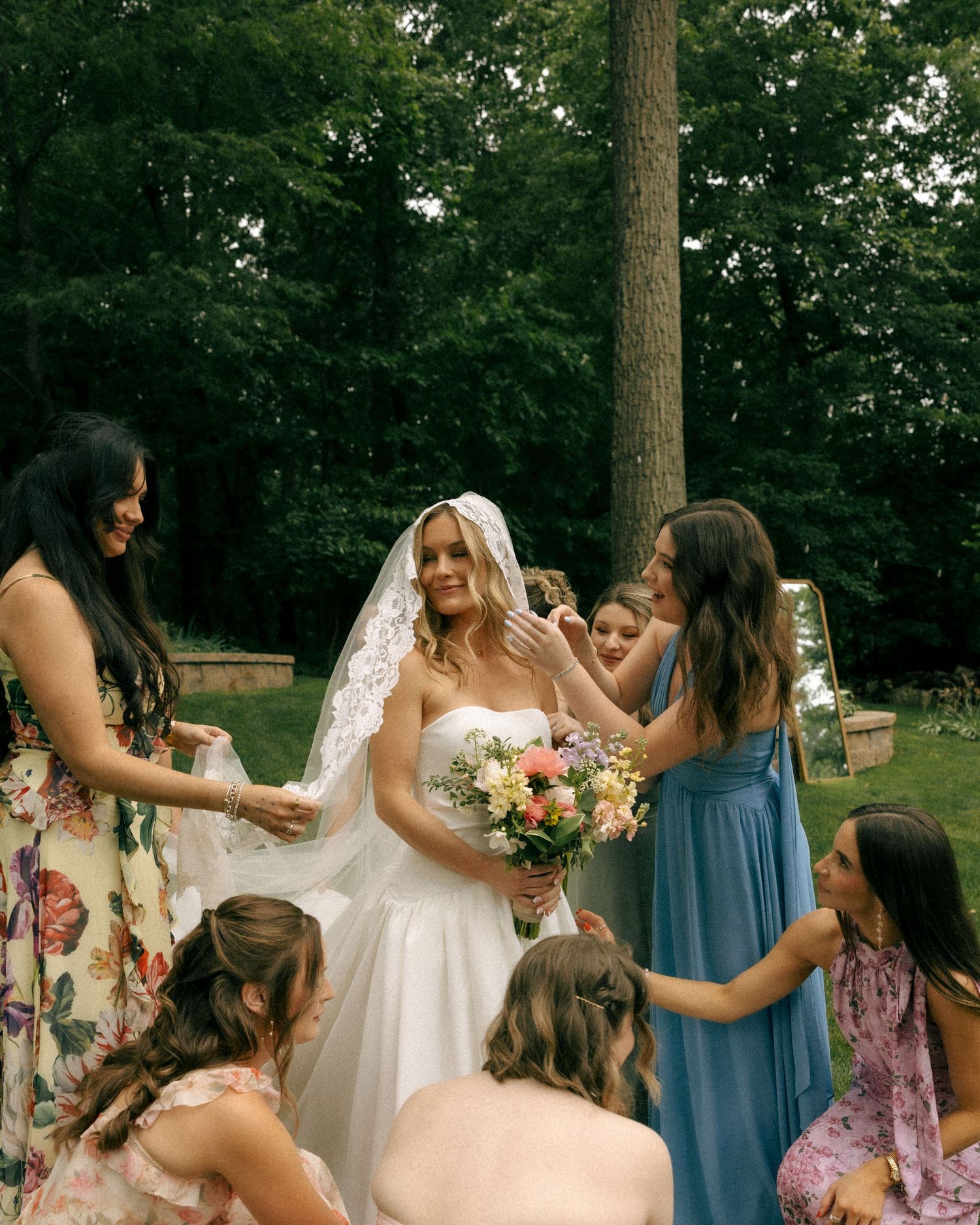 Bride in white gown and veil holding bouquet surrounded by bridesmaids in floral and blue dresses outdoors.
