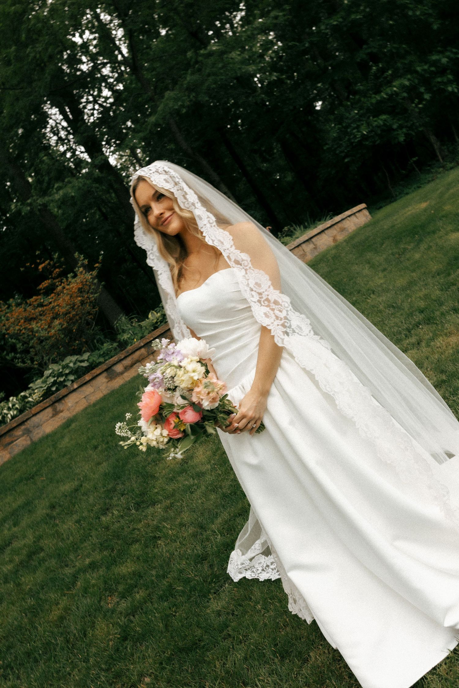 Bride in white gown and lace veil holding colorful bouquet, standing on green lawn with trees in background.