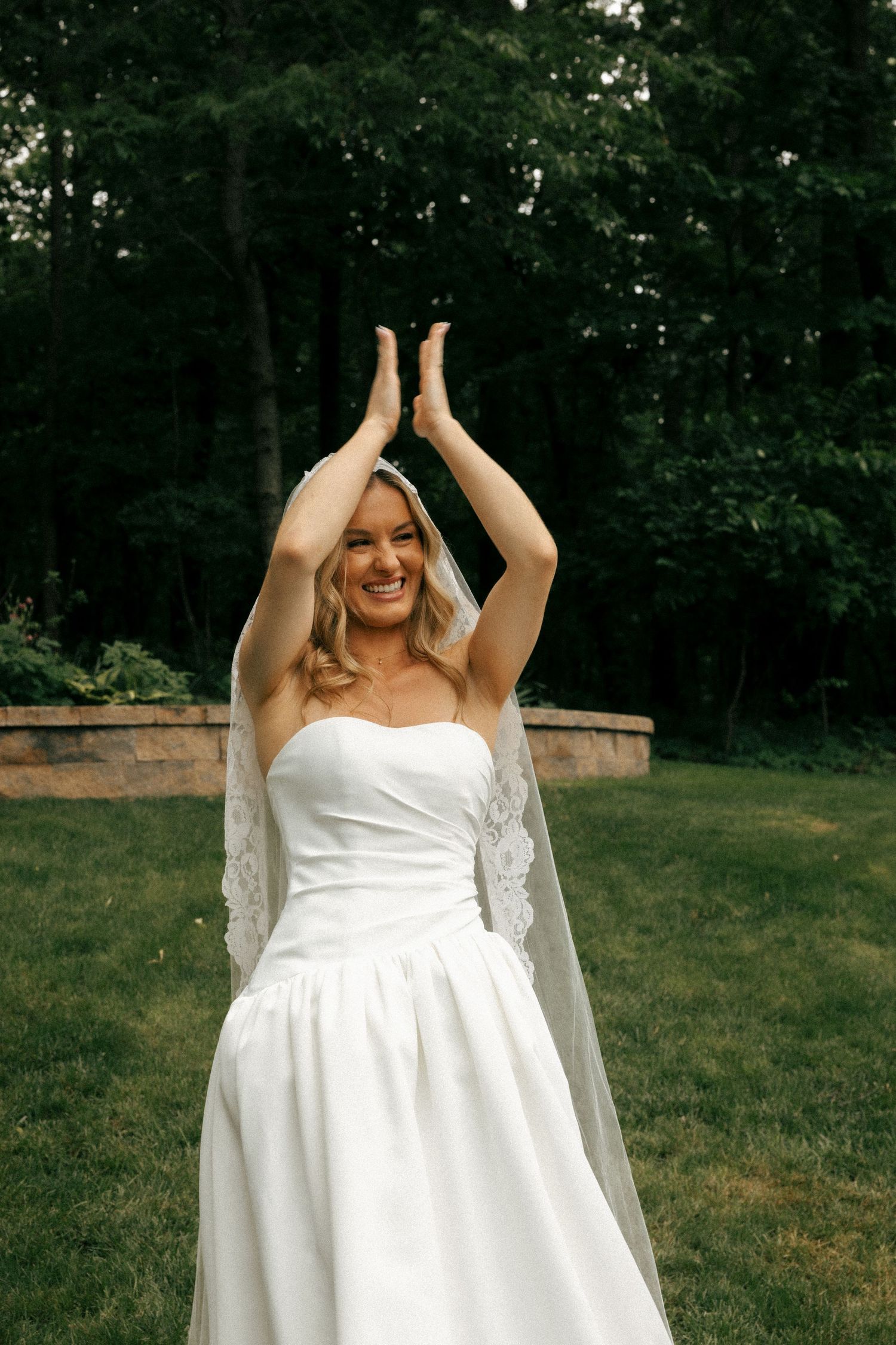 Smiling bride in strapless white gown and lace veil raises hands overhead outdoors on green lawn.