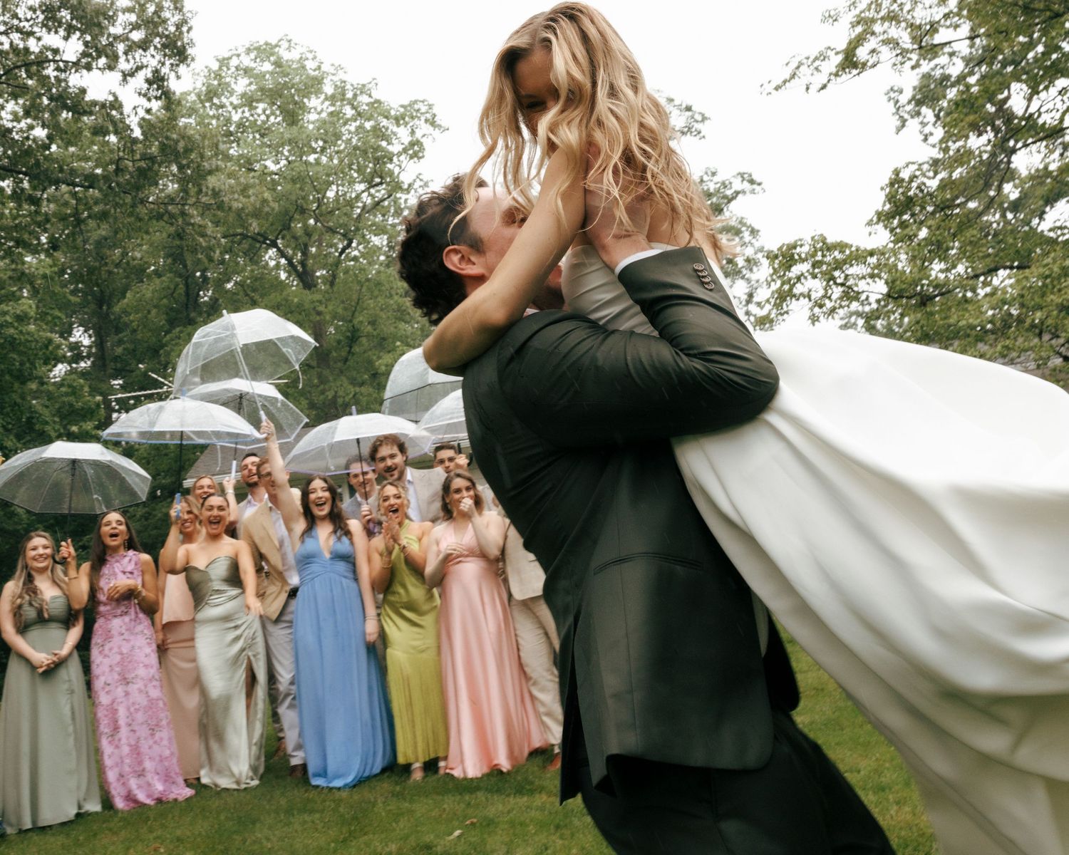 Groom lifts bride joyfully outdoors as wedding guests in colorful gowns celebrate with umbrellas on a rainy day.