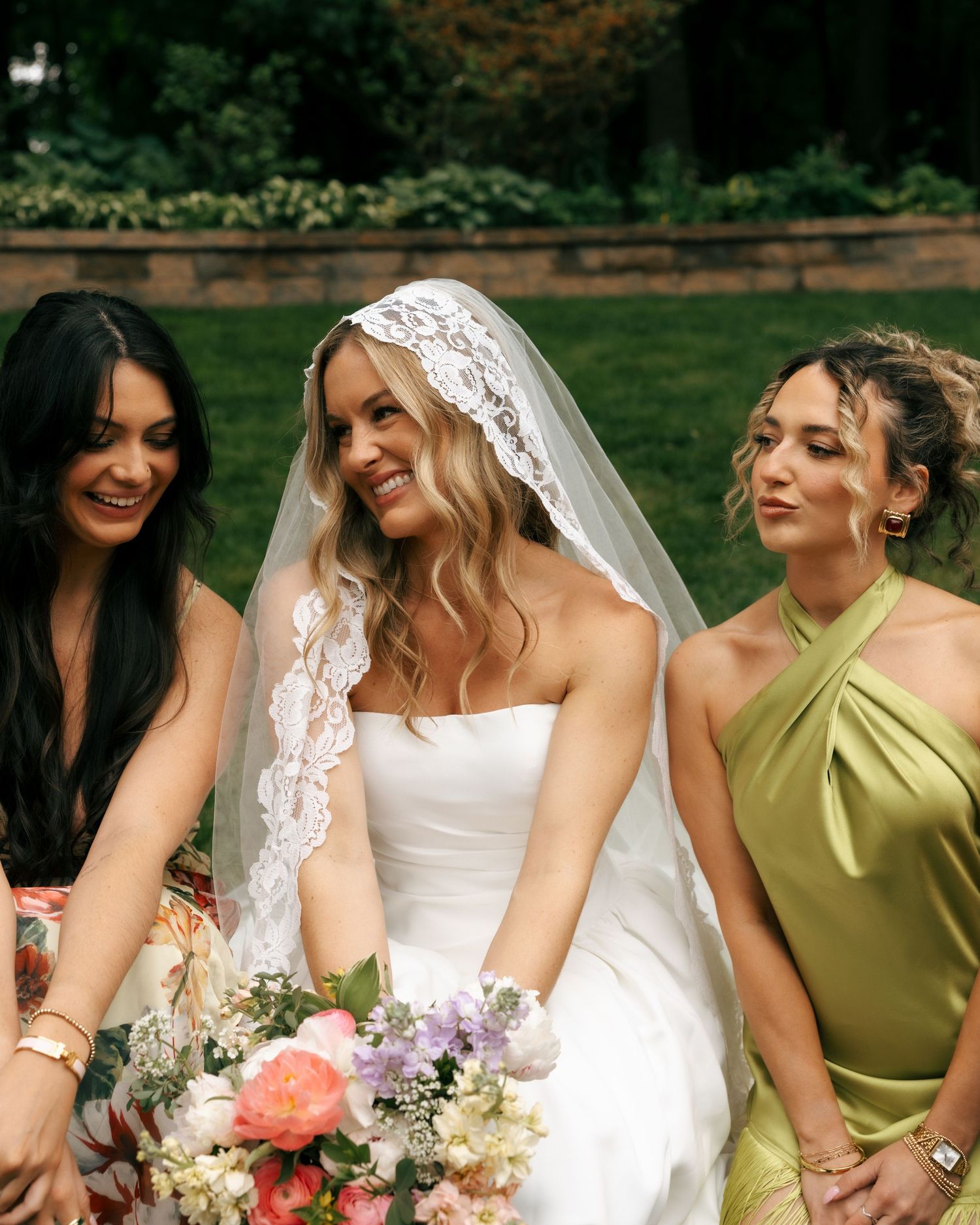 Bride in white gown and lace veil holds floral bouquet, smiling with two bridesmaids outdoors.