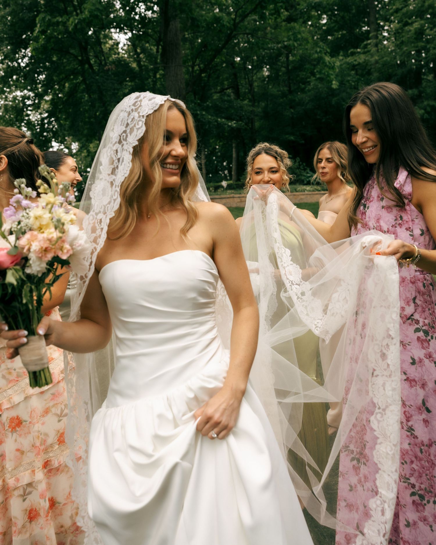 Smiling bride in white strapless gown and lace veil holds bouquet while bridesmaids in floral dresses assist outdoors.