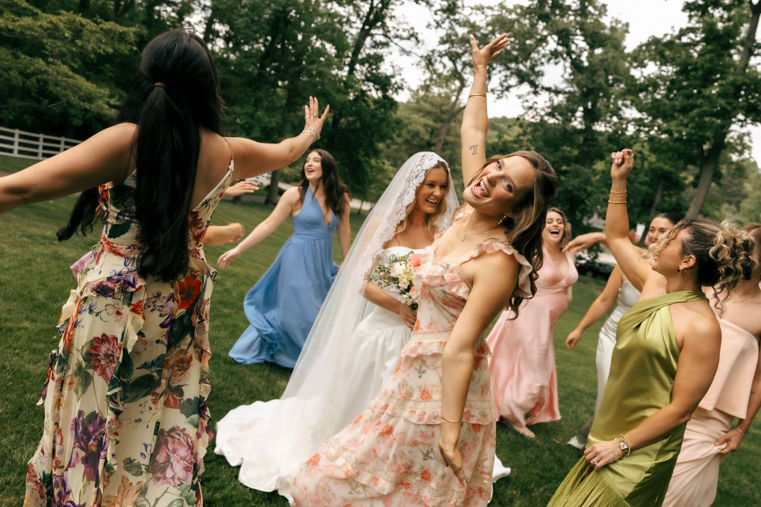Bride in floral gown dances joyfully with bridesmaids in colorful dresses outdoors at a wedding celebration.