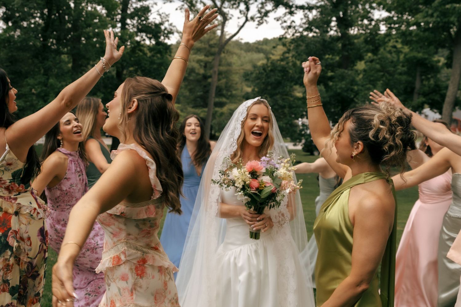 Bride in white gown and veil laughs joyfully surrounded by cheering bridesmaids in colorful dresses outdoors.