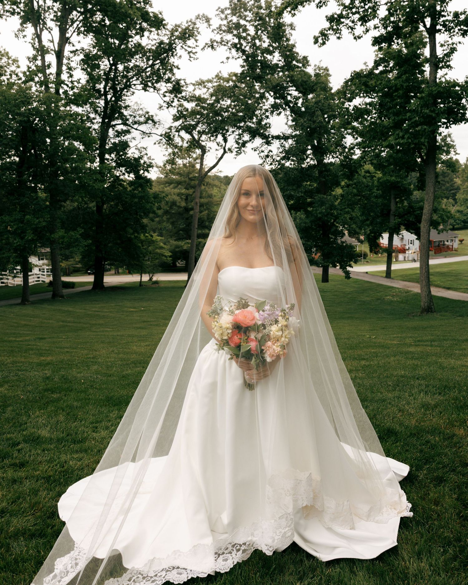 Bride in strapless white ballgown with lace-trimmed cathedral veil holding colorful bouquet on green lawn with trees.