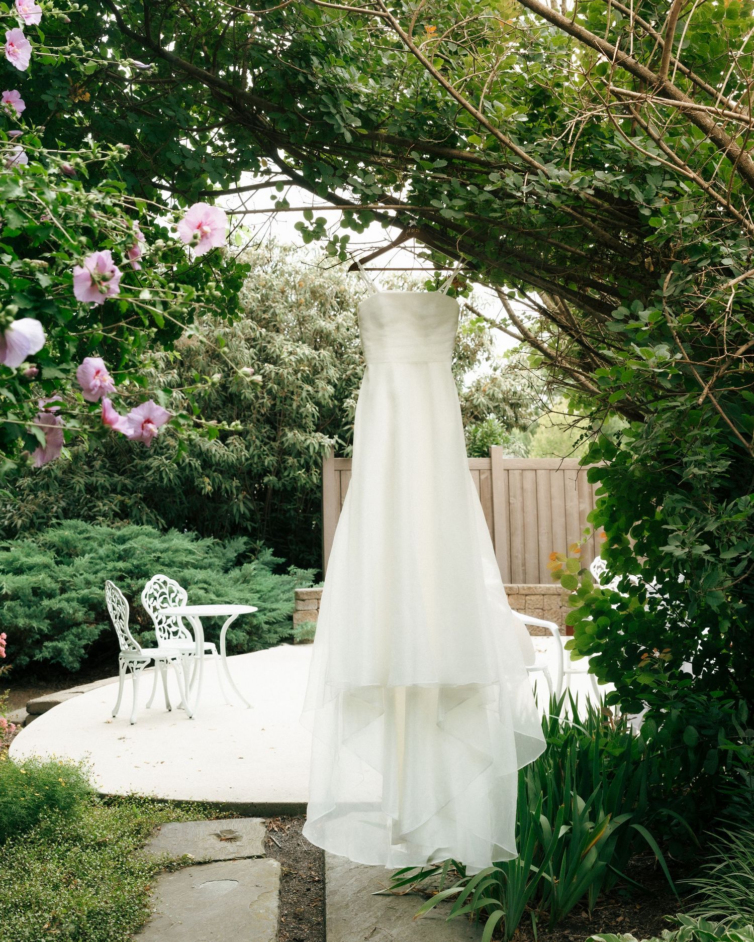White wedding dress hanging from a tree branch in a lush garden with white patio furniture and pink flowers.