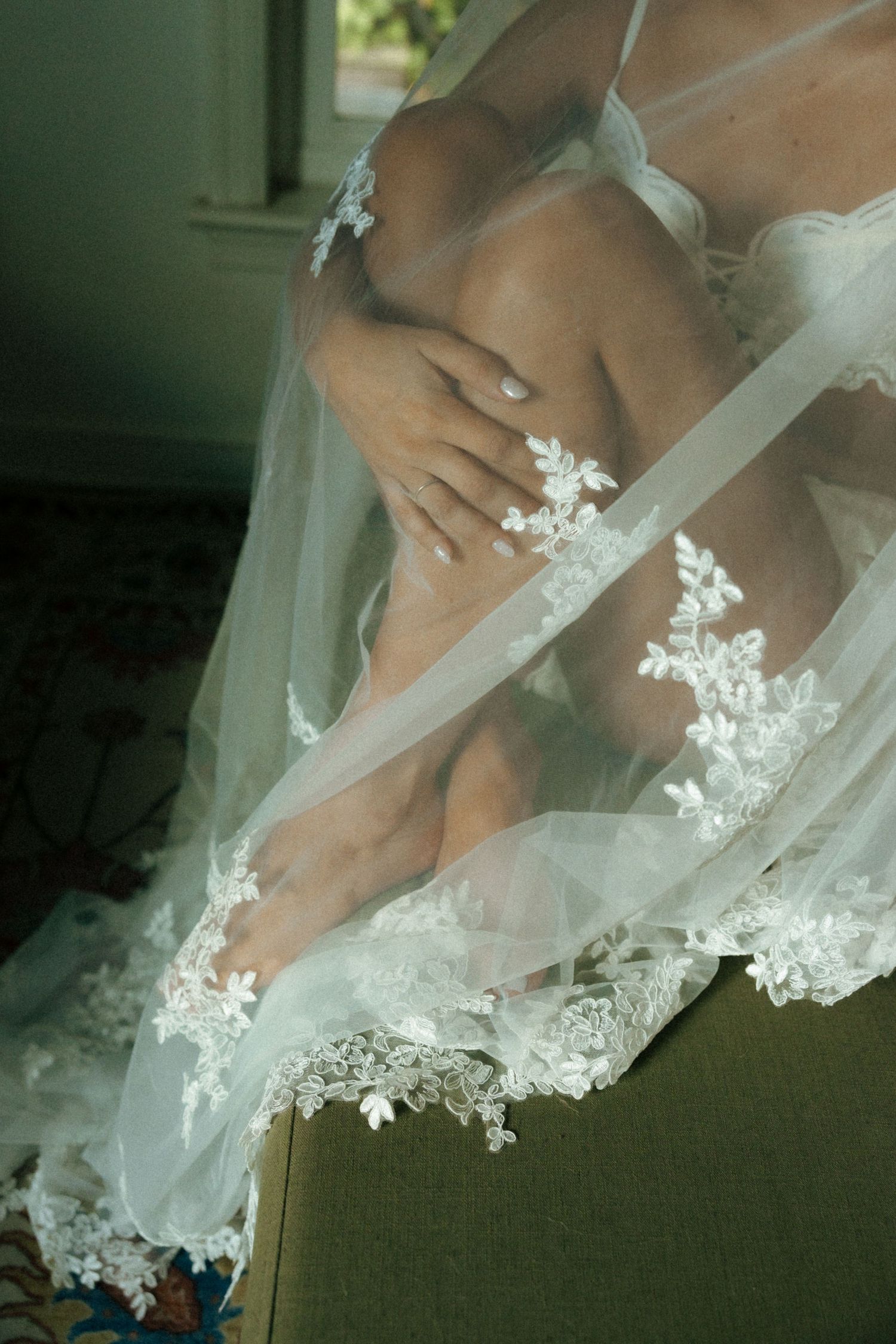 Bride sitting in chair draped in delicate white lace wedding veil with floral embroidery details.