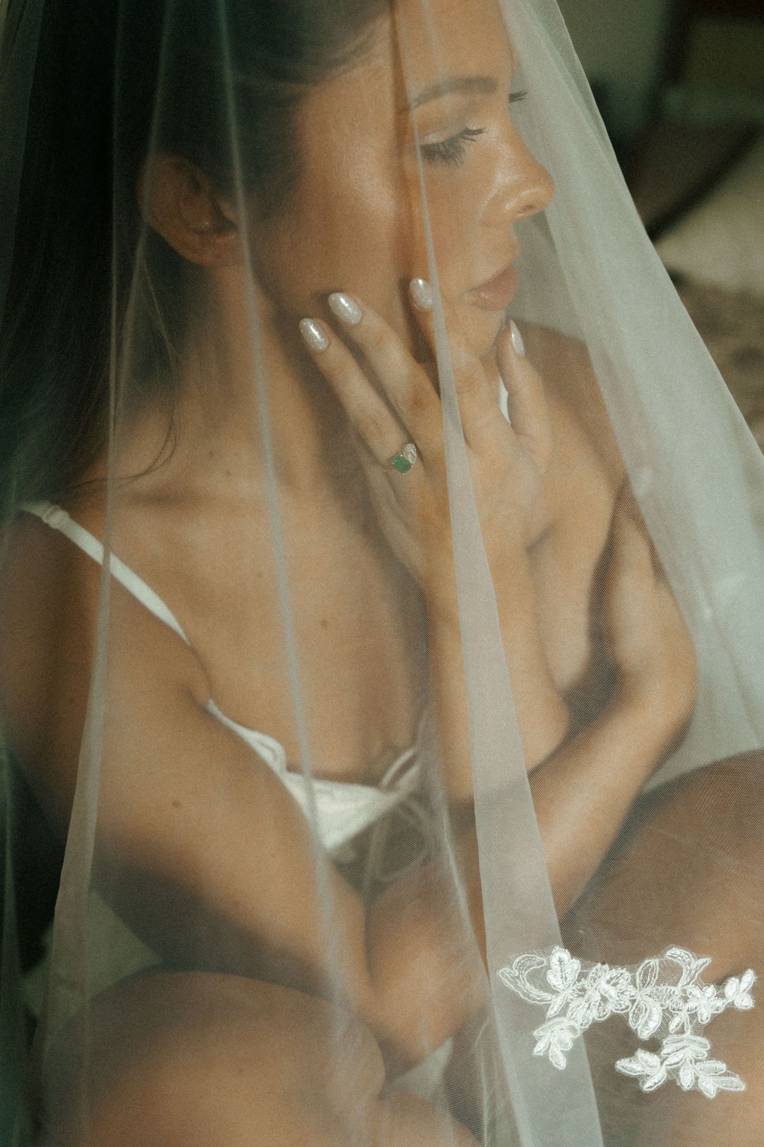 Bride in white lingerie draped under a sheer lace veil, posing elegantly with manicured nails and a delicate ring.