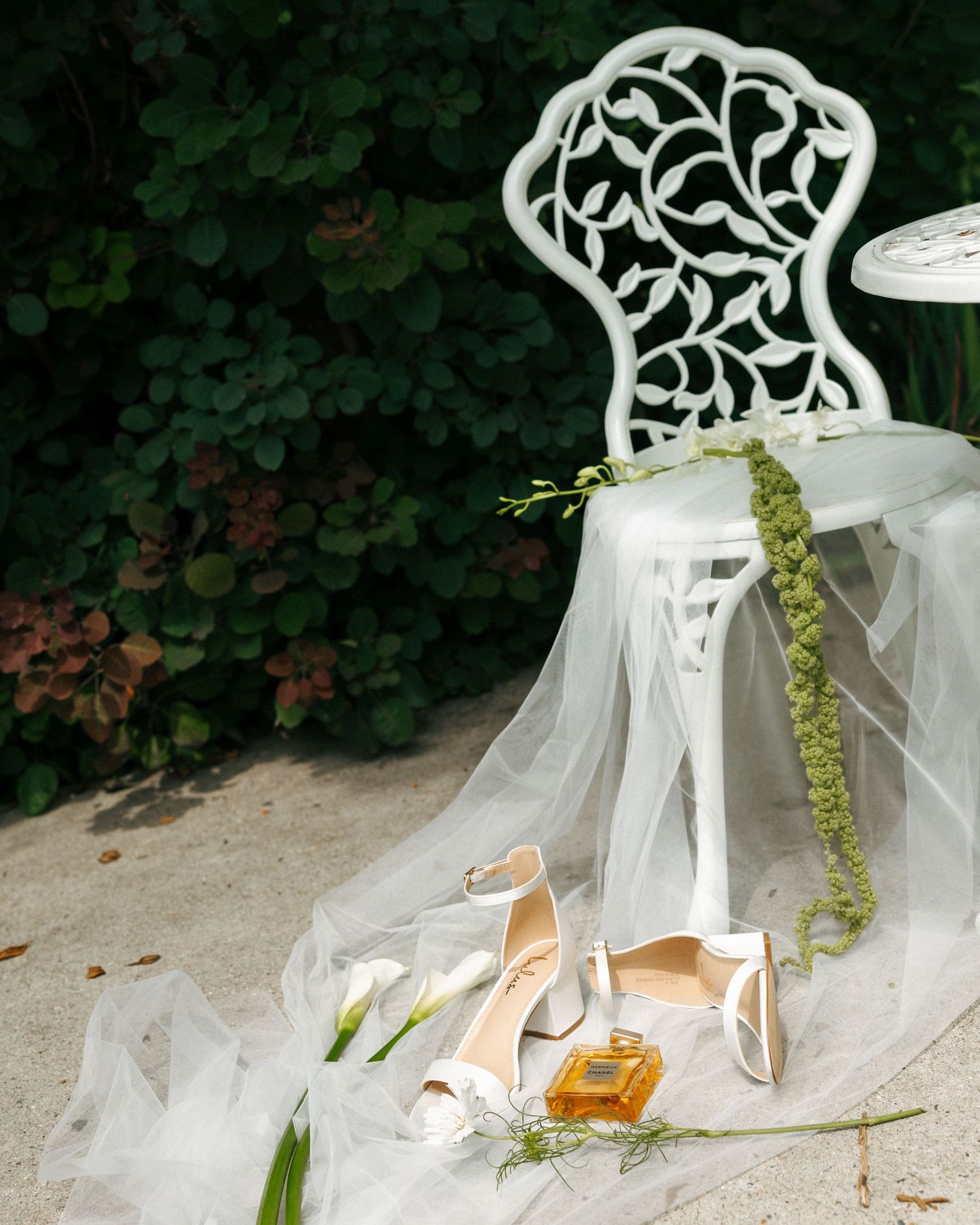 White bridal heels and perfume bottle on tulle veil beside ornate white garden chair with greenery backdrop.