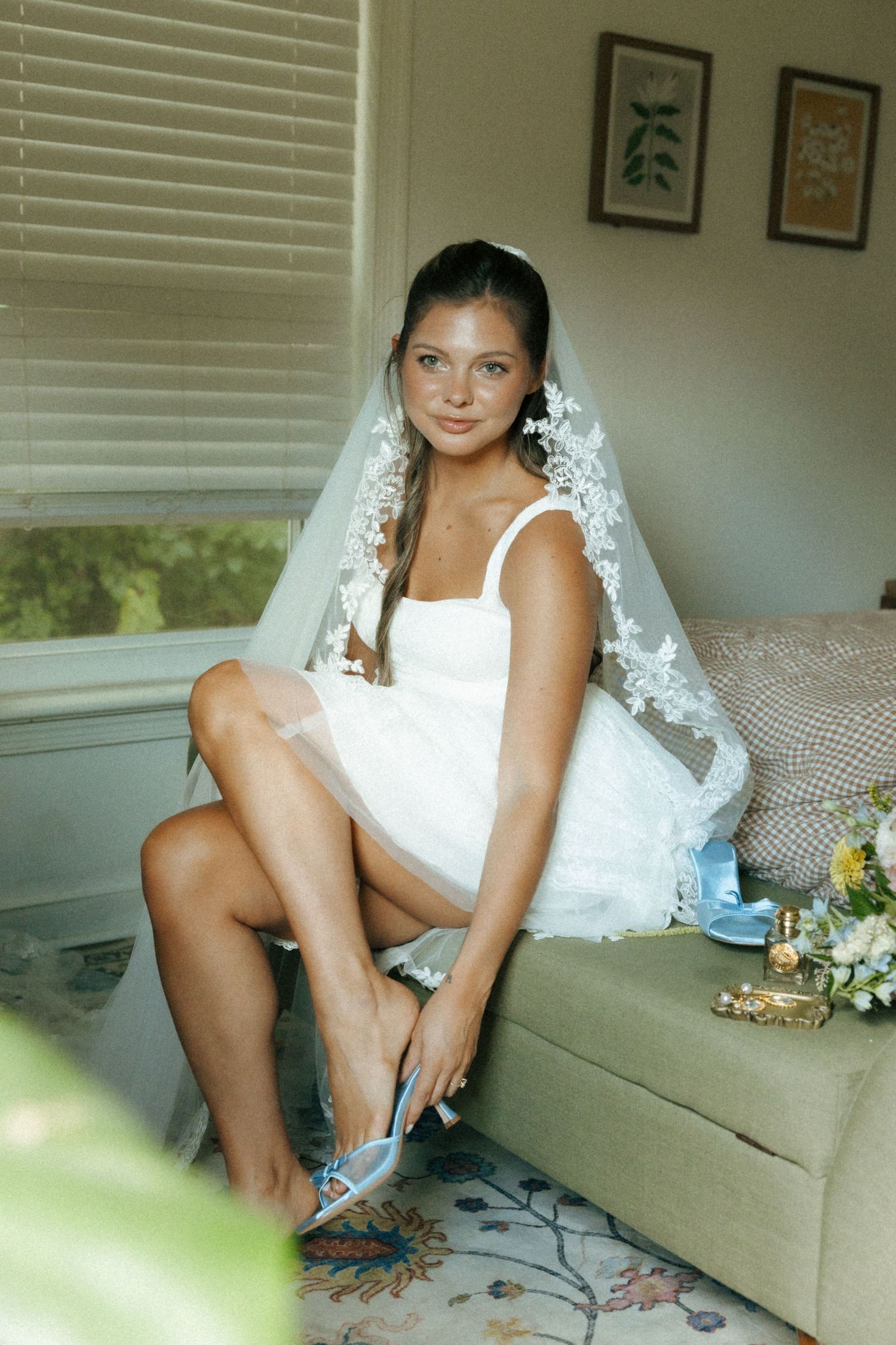 Bride in short white dress and lace veil sits on sofa adjusting blue heels near window on wedding day.
