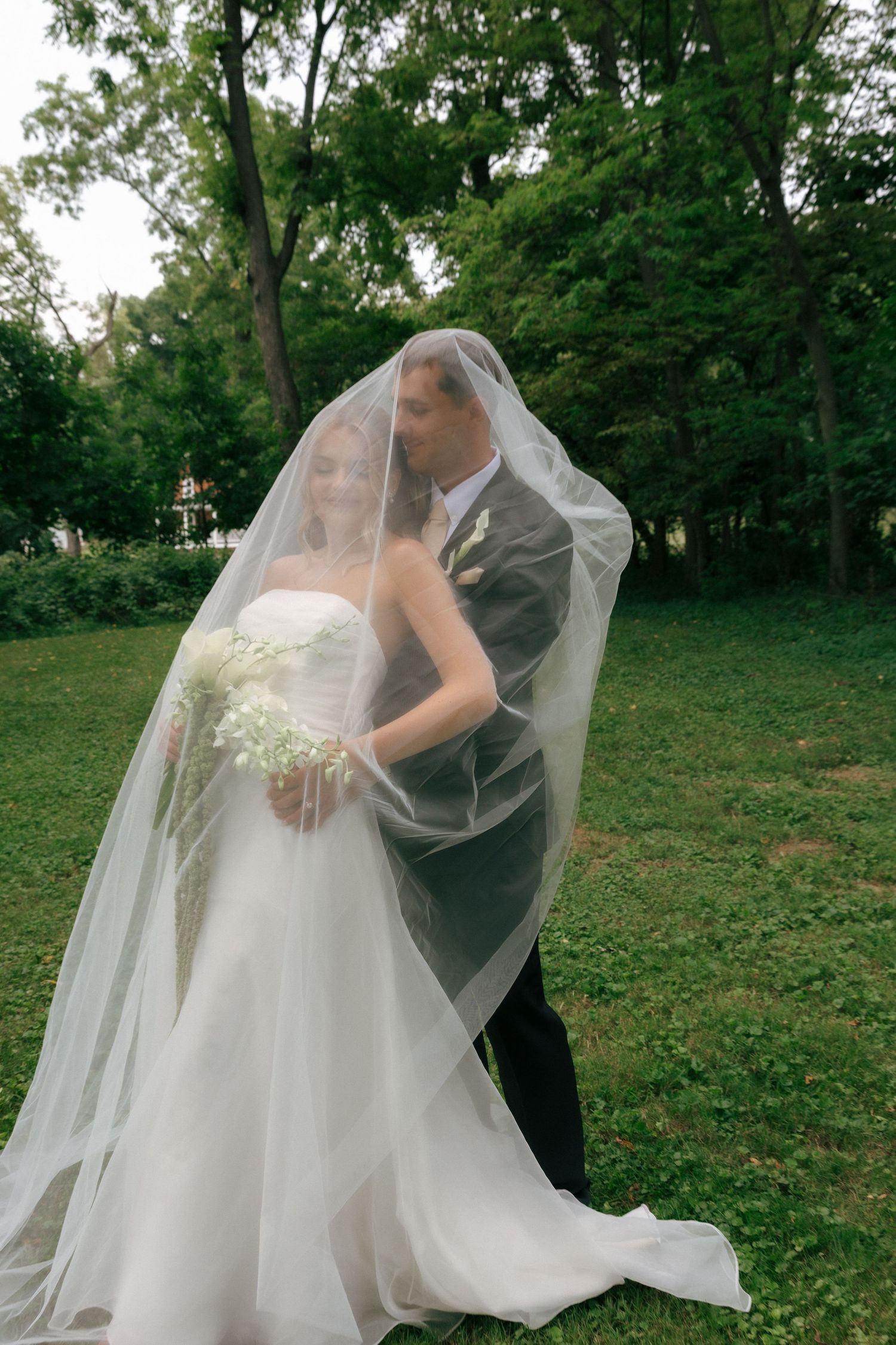 Bride and groom share romantic moment under flowing white veil in lush green park setting on wedding day.