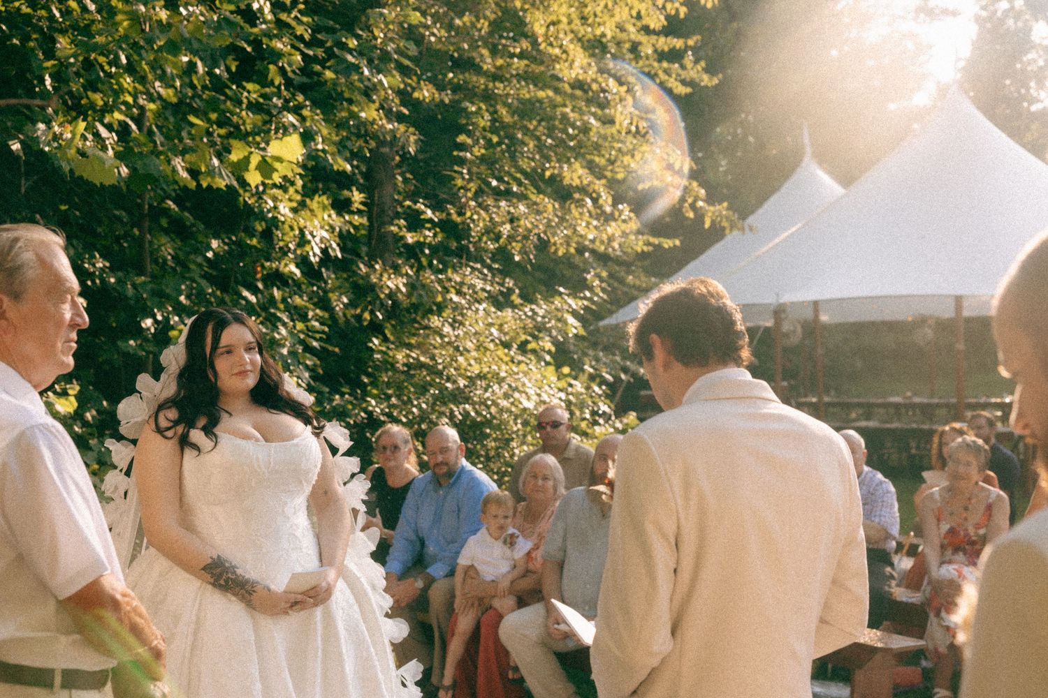Outdoor wedding ceremony with bride in white gown facing groom, guests seated under sunlit trees near white tent.